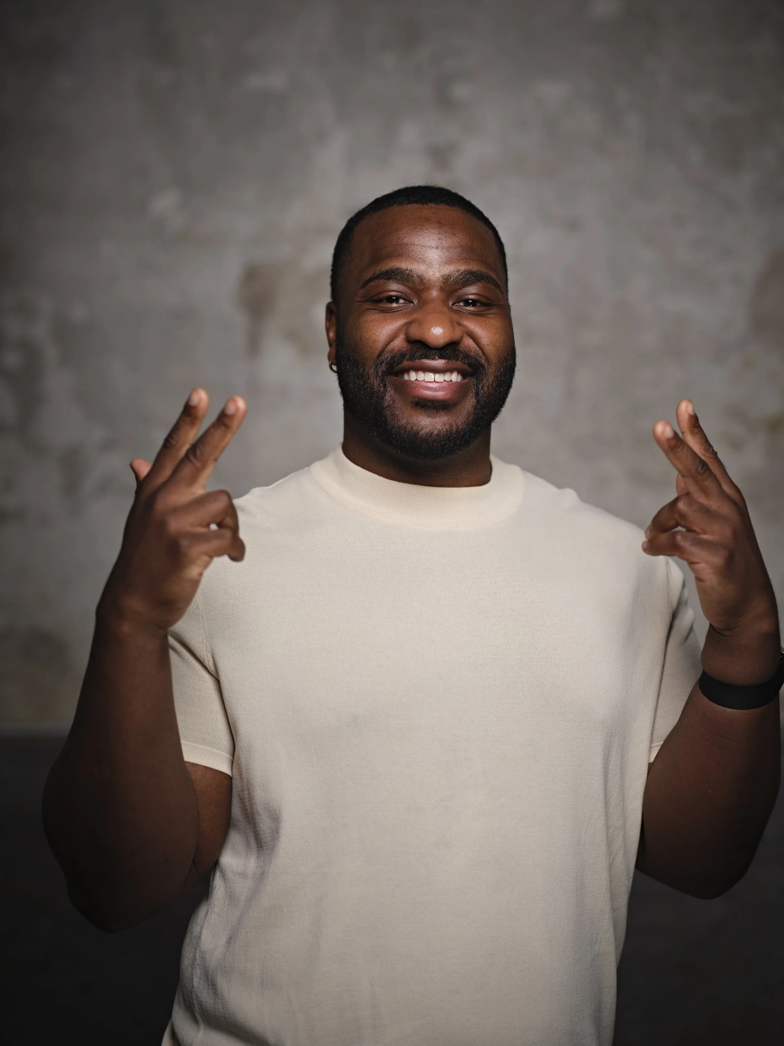 Smiling man with a beard making peace signs with both hands, wearing a beige T-shirt against a textured gray background.