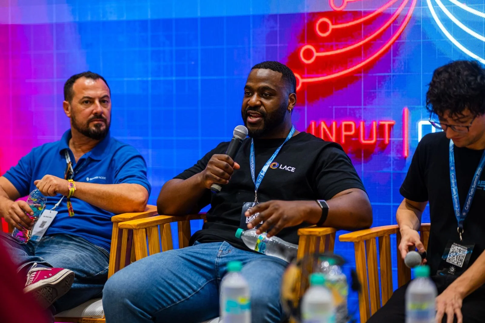 Three men sit on stage at a conference, with one speaking into a microphone. The man in the center is wearing a black T-shirt with a Lace logo, while the other two men wear blue and black T-shirts. Behind them is a large digital screen with colorful graphics and the word 'INPUT' visible.