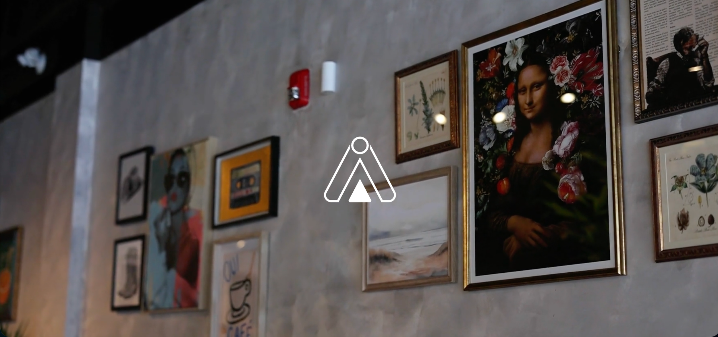 A wall decorated with various framed pictures, including a large painting of the Mona Lisa surrounded by flowers, and smaller art pieces and text. There is a fire alarm and a white cylindrical object on the wall.