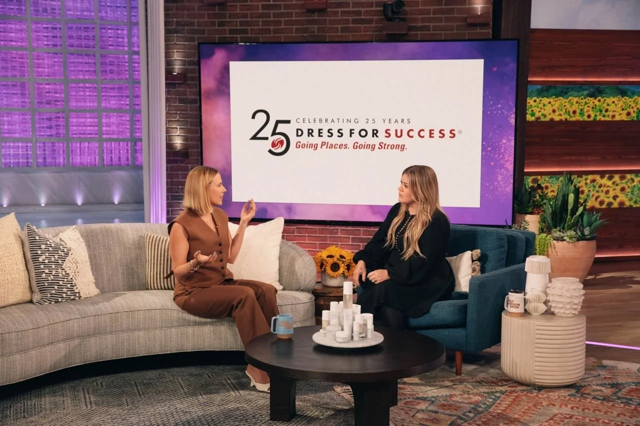 Two women are seated on sofas in a TV studio, engaging in a conversation. Behind them is a large screen displaying the logo and information for Dress for Success's 25th anniversary. The studio is decorated with pillows, plants, and a coffee table with various bottles and mugs.
