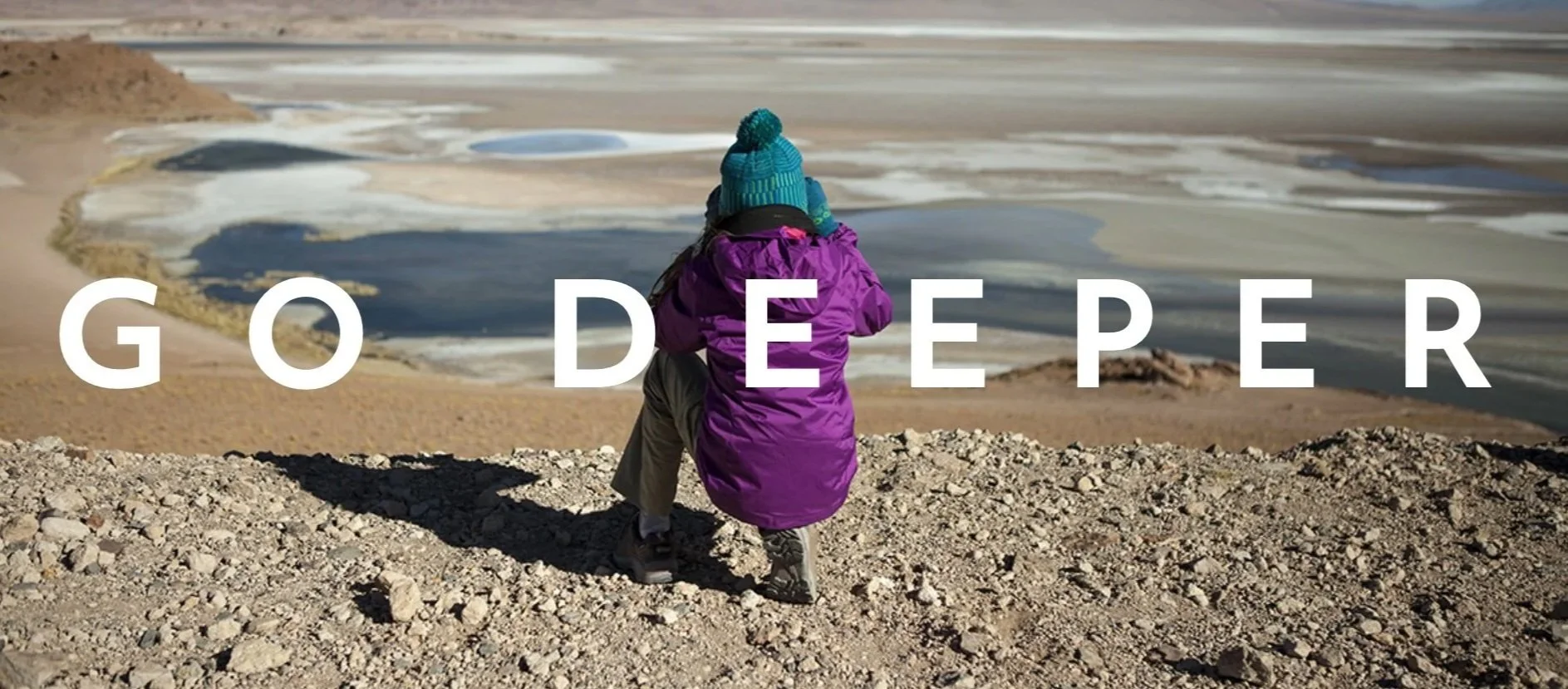 A person dressed in outdoor winter clothing sitting on a rocky hill overlooking a desert landscape with salt flats and shallow water. The words "GO DEEPER" are overlaid on the image.