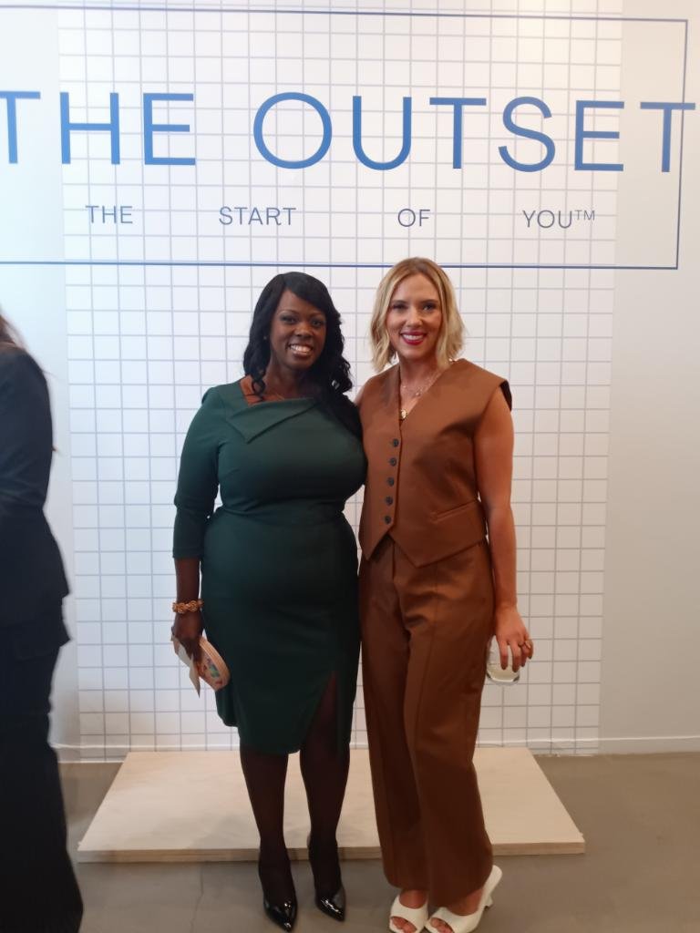 Two women standing together in front of a wall with the text 'THE OUTSET, THE START OF YOU.' One woman is wearing a dark green dress, and the other is wearing a brown sleeveless jumpsuit. They are smiling and posing for the photo.