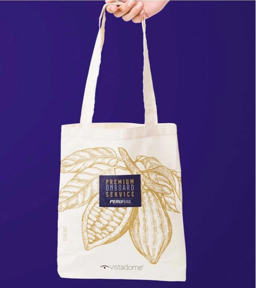 A person holding a white tote bag with gold illustrations of cocoa pods and leaves, with a black logo reading 'Premium Onboard Service Peru/Bail' against a blue background.
