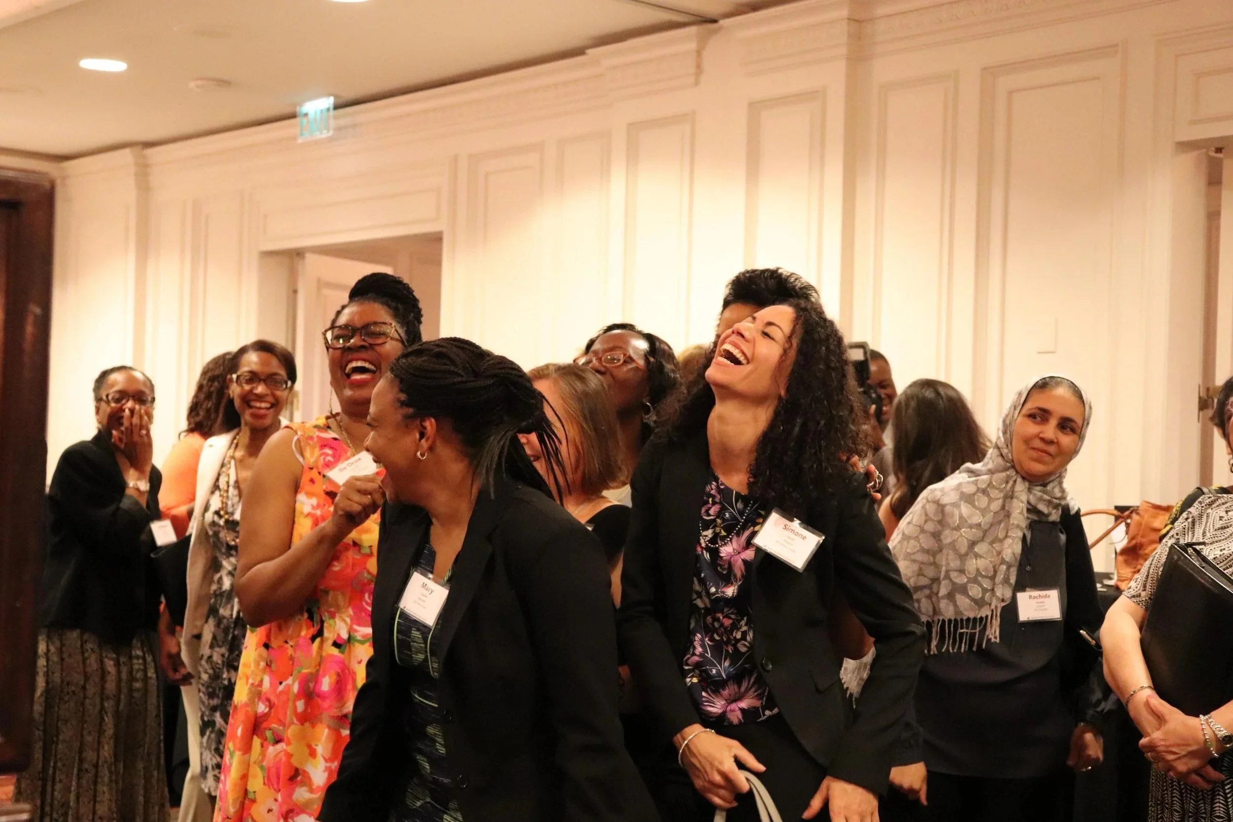 Group of women at a conference, laughing and smiling, wearing name tags, in a room with cream-colored walls and warm lighting.