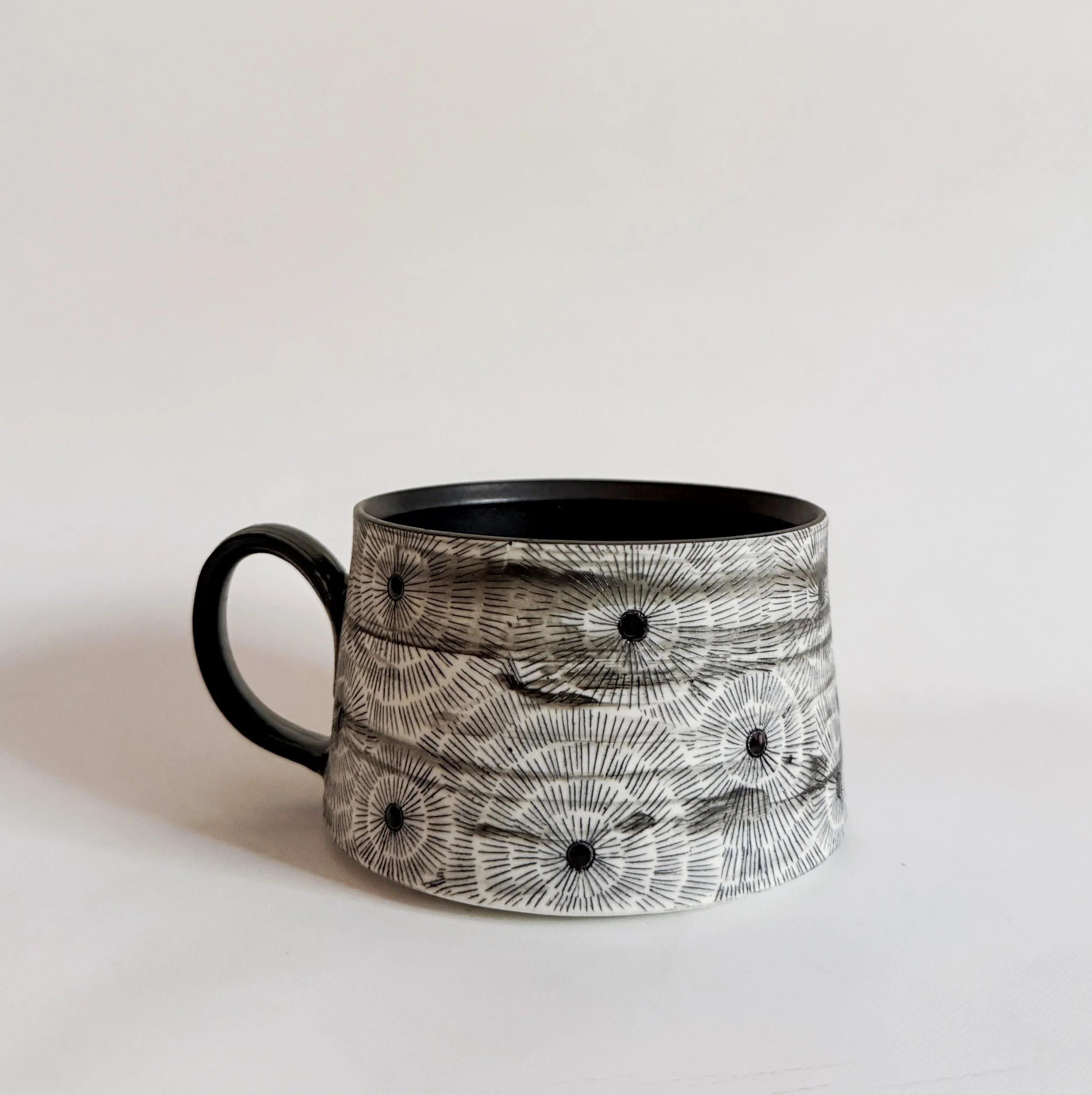 A white mug with black intricate radial line patterns and black dots, set against a plain white background.