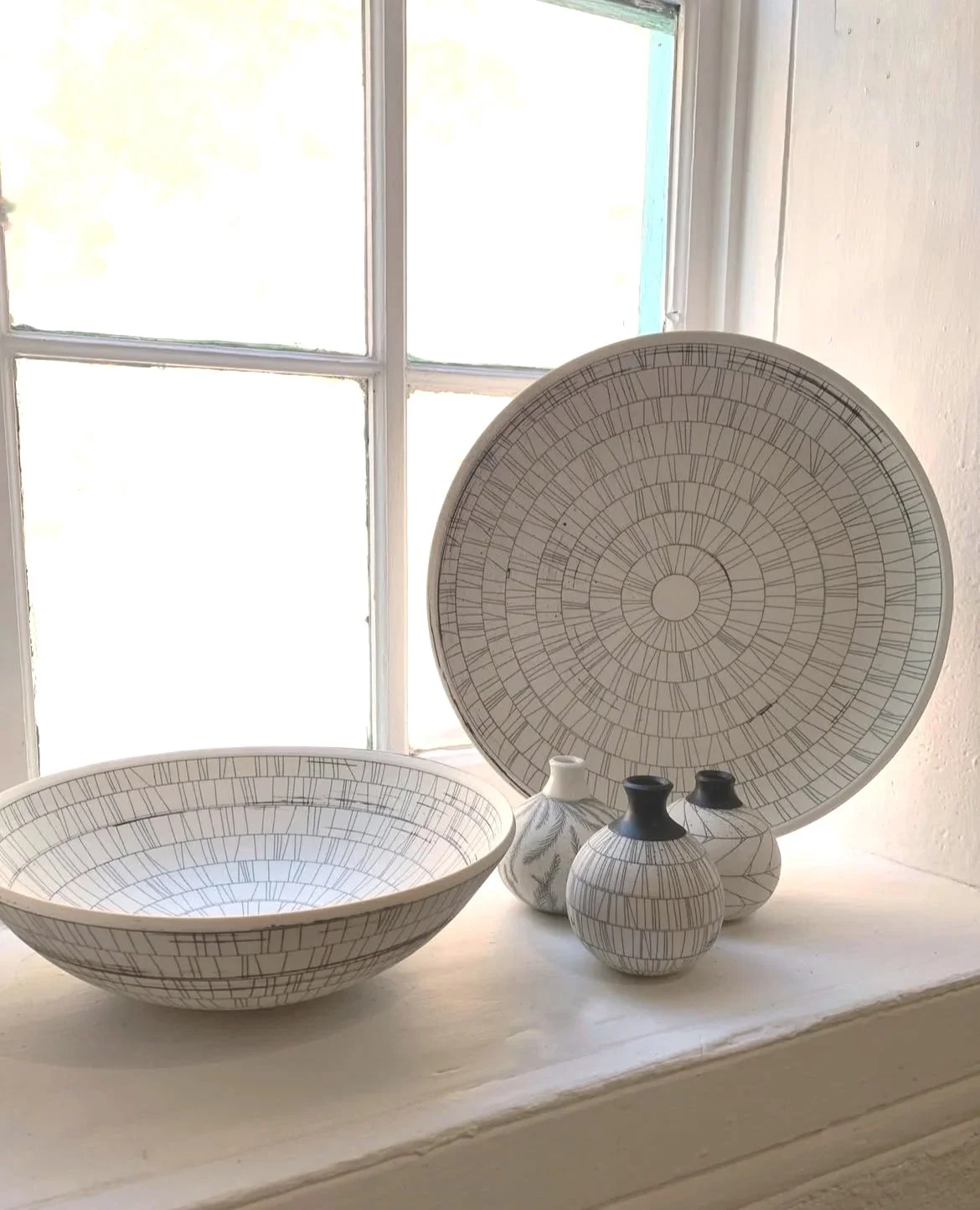A ceramic bowl and three vases with black lids, all with coordinating geometric line patterns, placed on a white windowsill in front of a window with white framing.