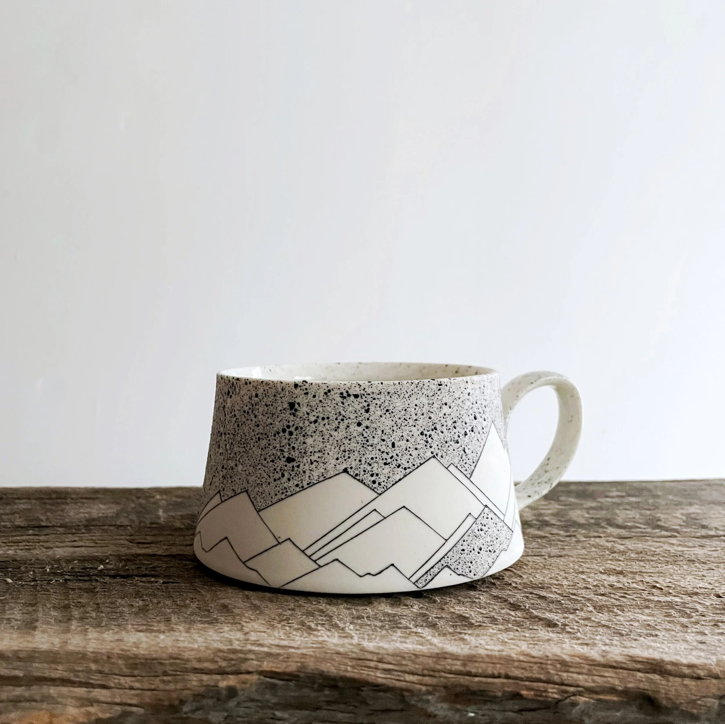 A ceramic mug with a black and white mountain design on a wooden surface against a plain white background.