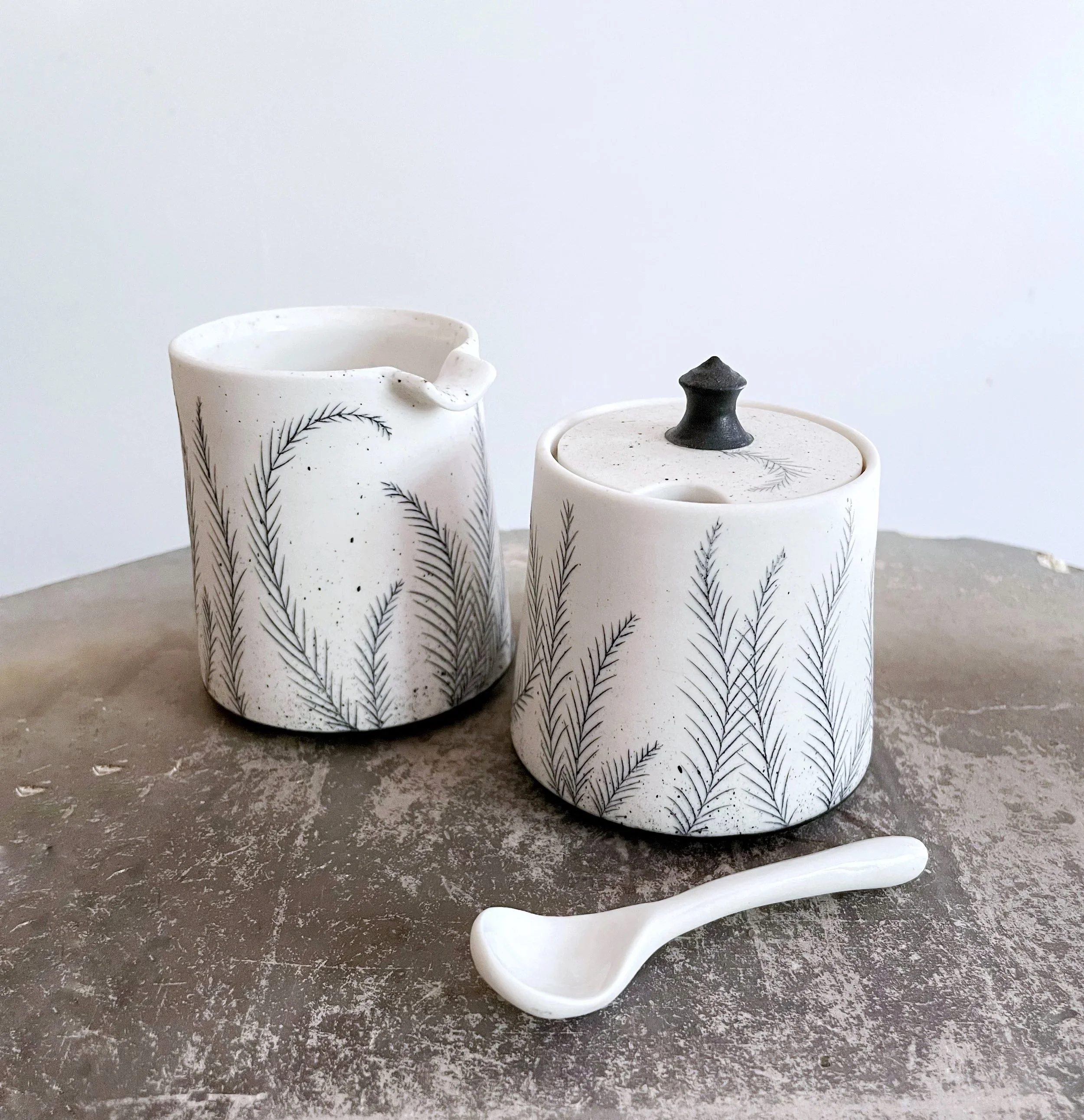 Two white ceramic containers with black pine branch designs, one with a lid and one with a spout, and a small ceramic spoon on a textured gray surface.