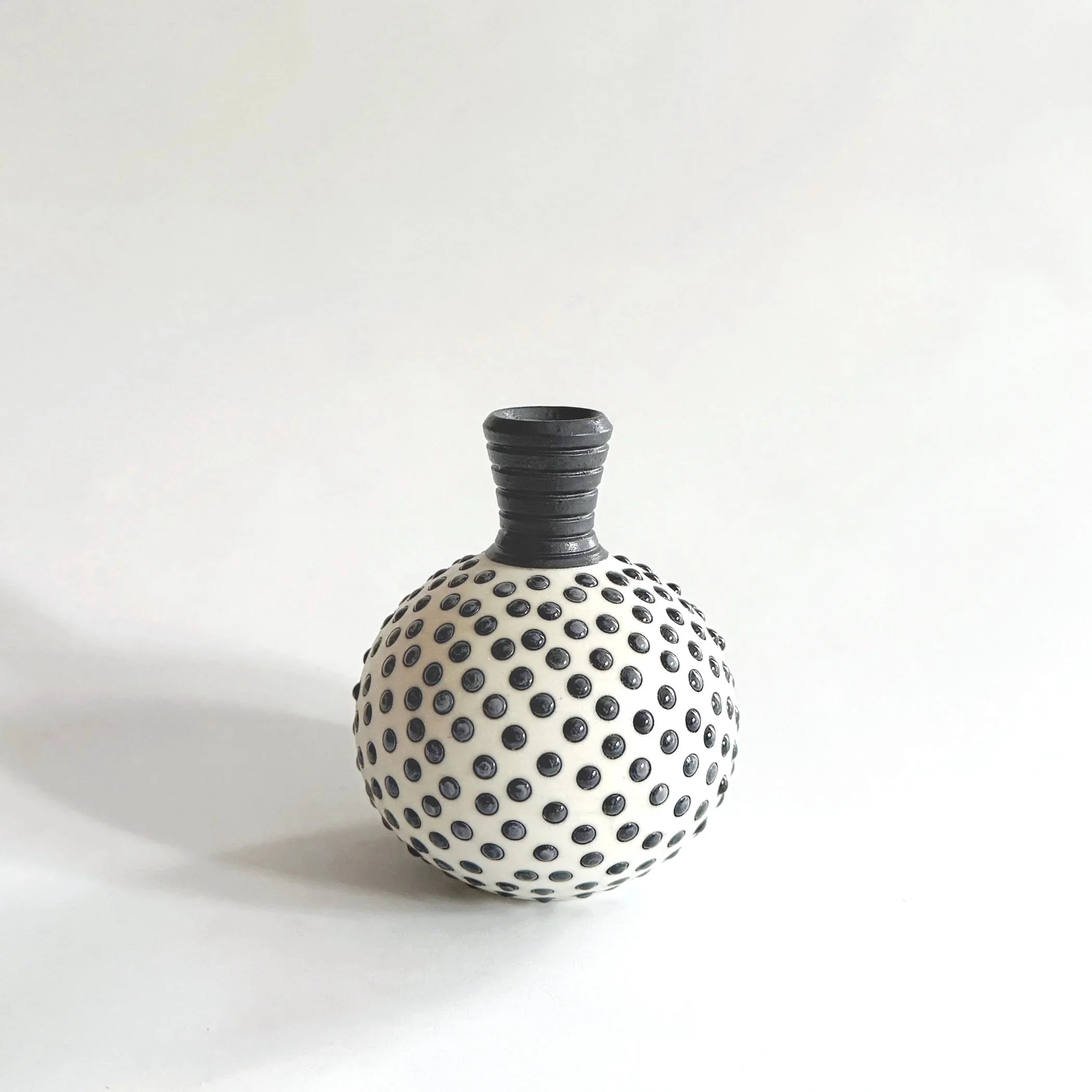 A decorative ceramic vase with a round body covered in black dots and a short, ribbed black neck, placed on a plain white background.