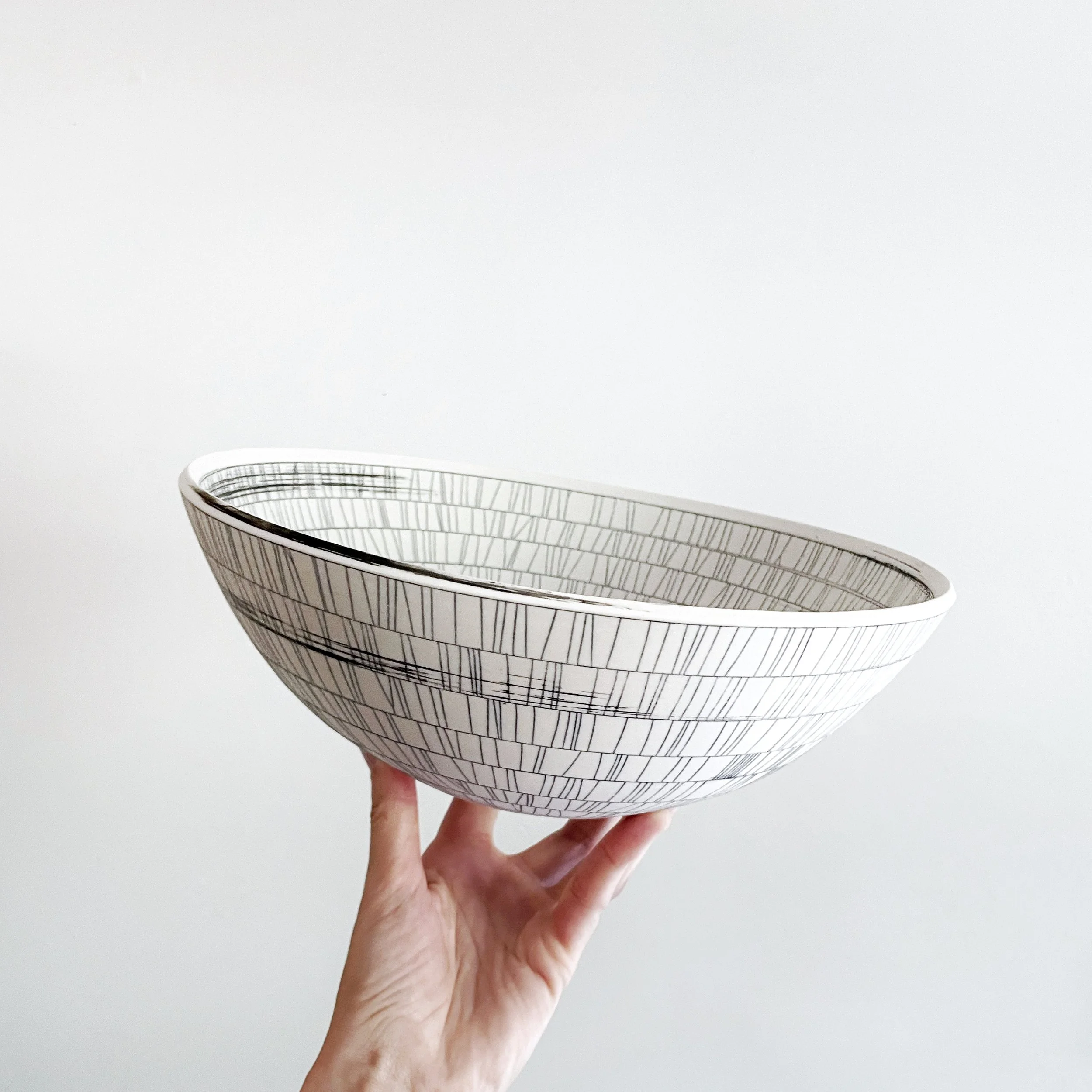 Hand holding a white ceramic bowl with black linear geometric pattern against a plain background.