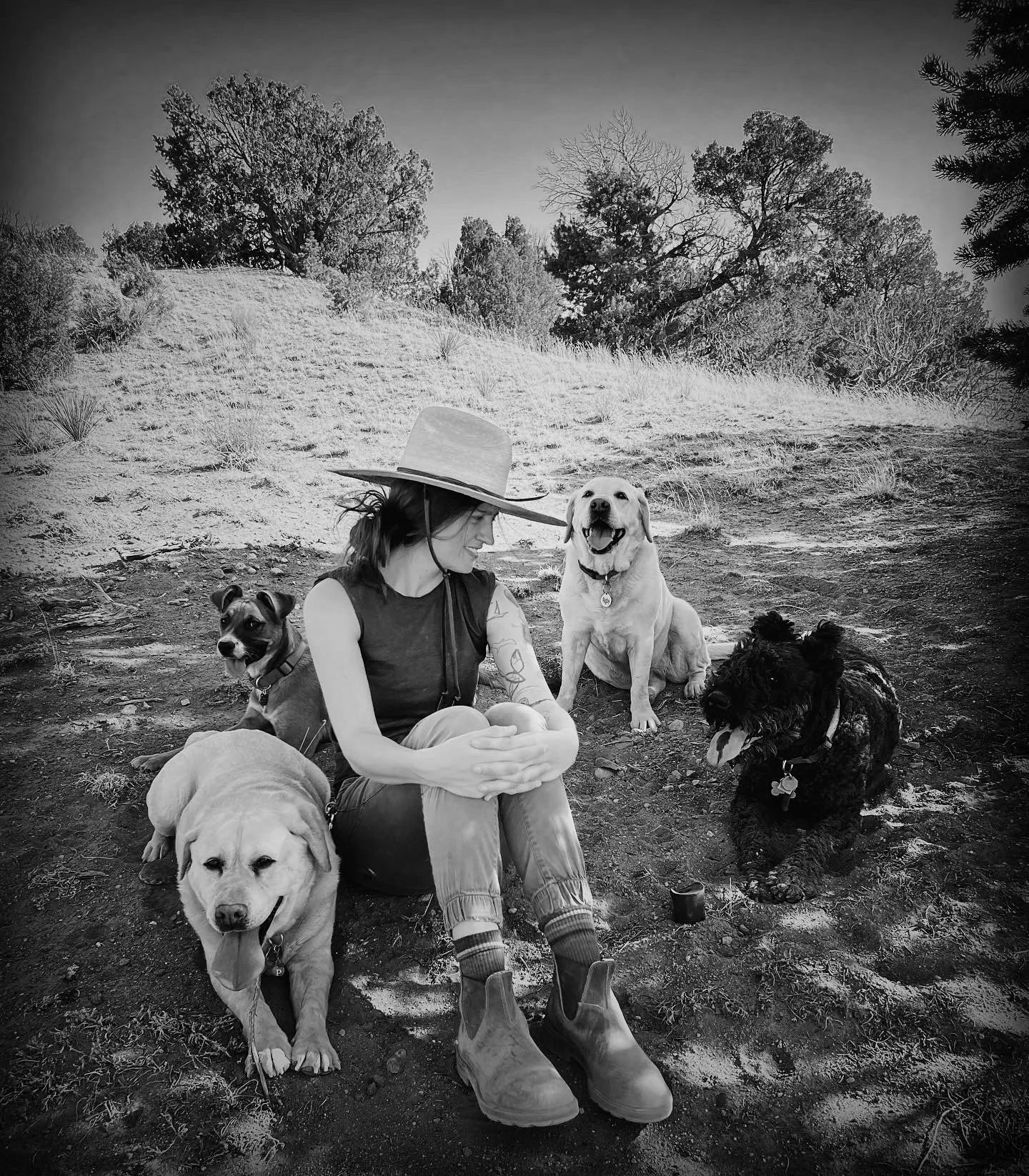 A woman sitting on the ground with four dogs around her in an outdoor natural setting with trees and grass in the background. She is wearing a wide-brimmed hat and casual clothing.