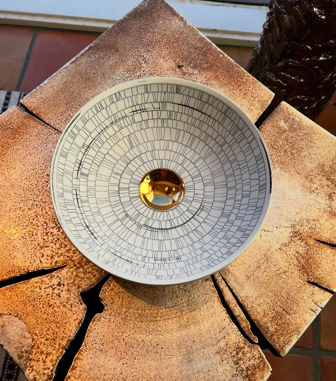 A decorative bowl with a white and black geometric pattern and a reflective golden center, placed on a cracked, peach-colored tiled surface.