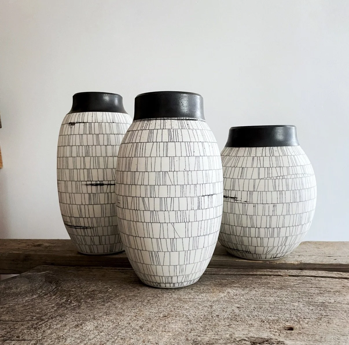 Three white ceramic vases with black tops and a geometric black line pattern, placed on a rustic wooden surface against a plain white wall.