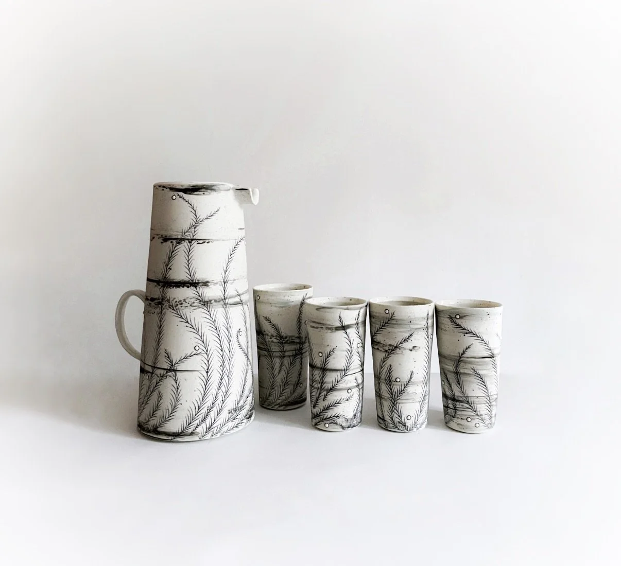 Ceramic pitcher and four ceramic tumblers with black leaf and spiral design on a white background.