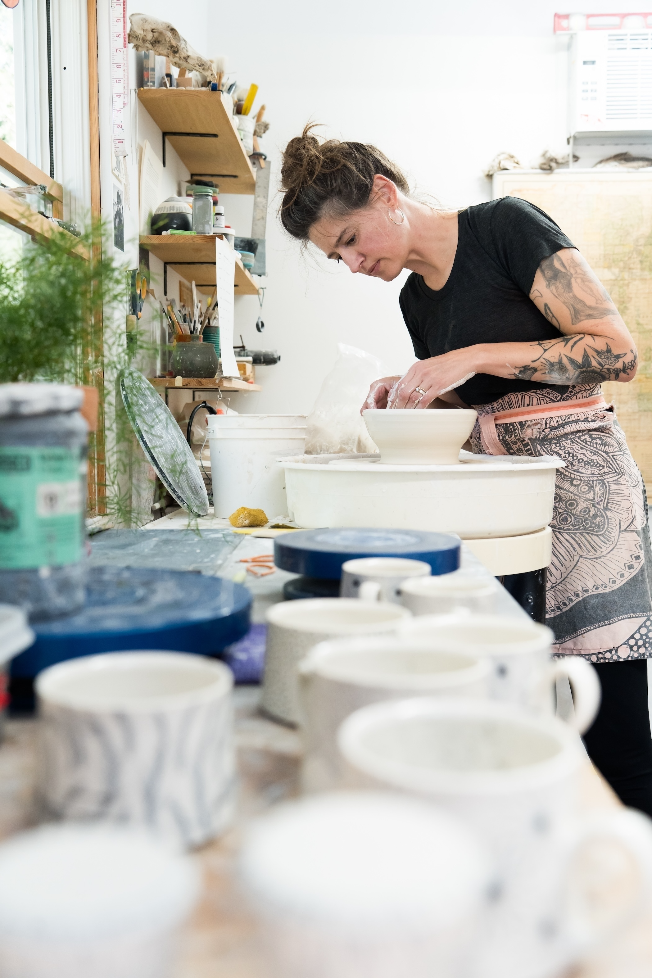 A woman with tattoos wearing a black t-shirt and a patterned apron is working on pottery at a pottery studio. She is shaping a bowl on a pottery wheel with various cups and supplies on the workbench in front of her.