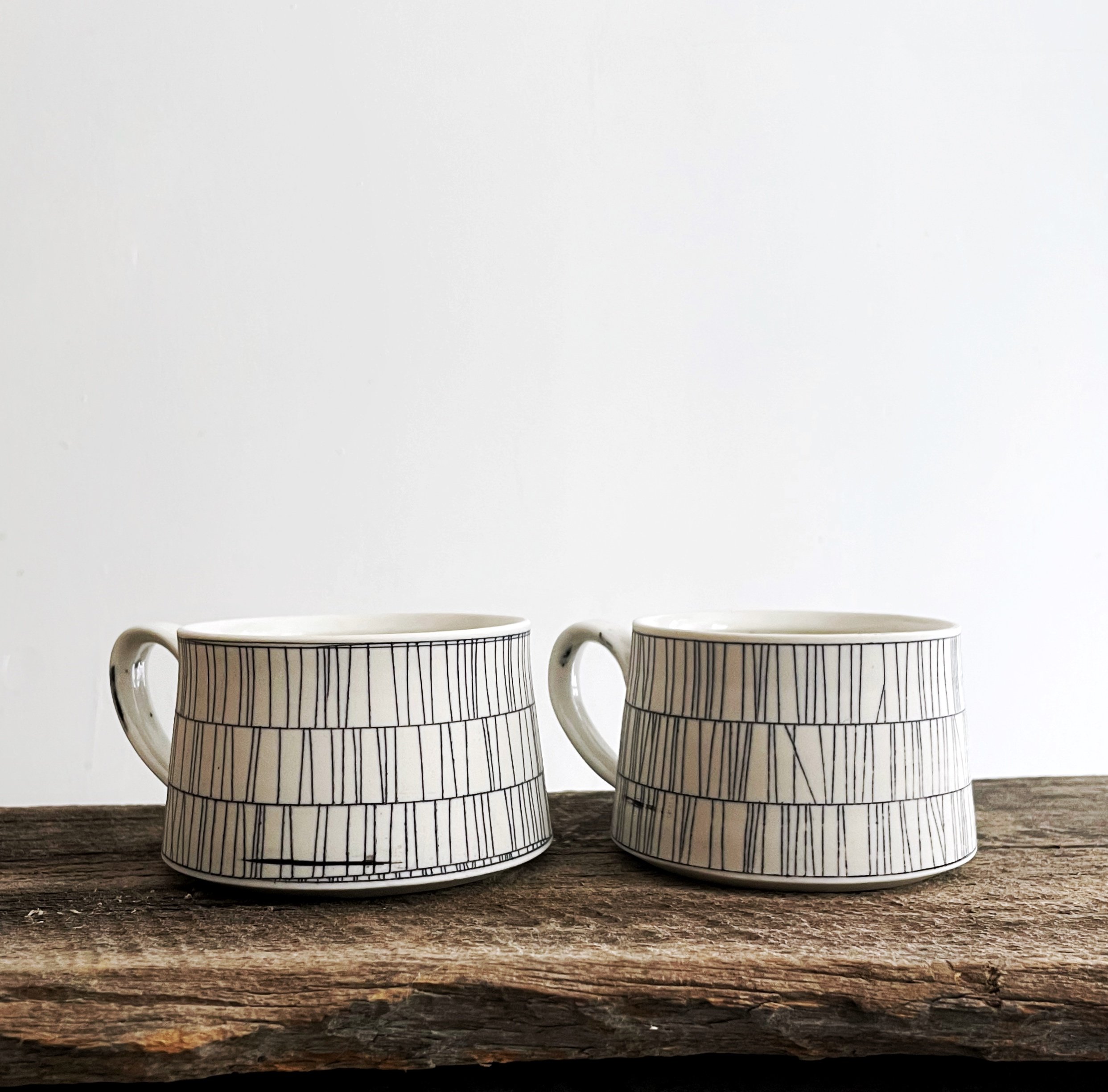 Pair of white ceramic mugs with black line patterns on a rustic wooden surface against a plain white wall.