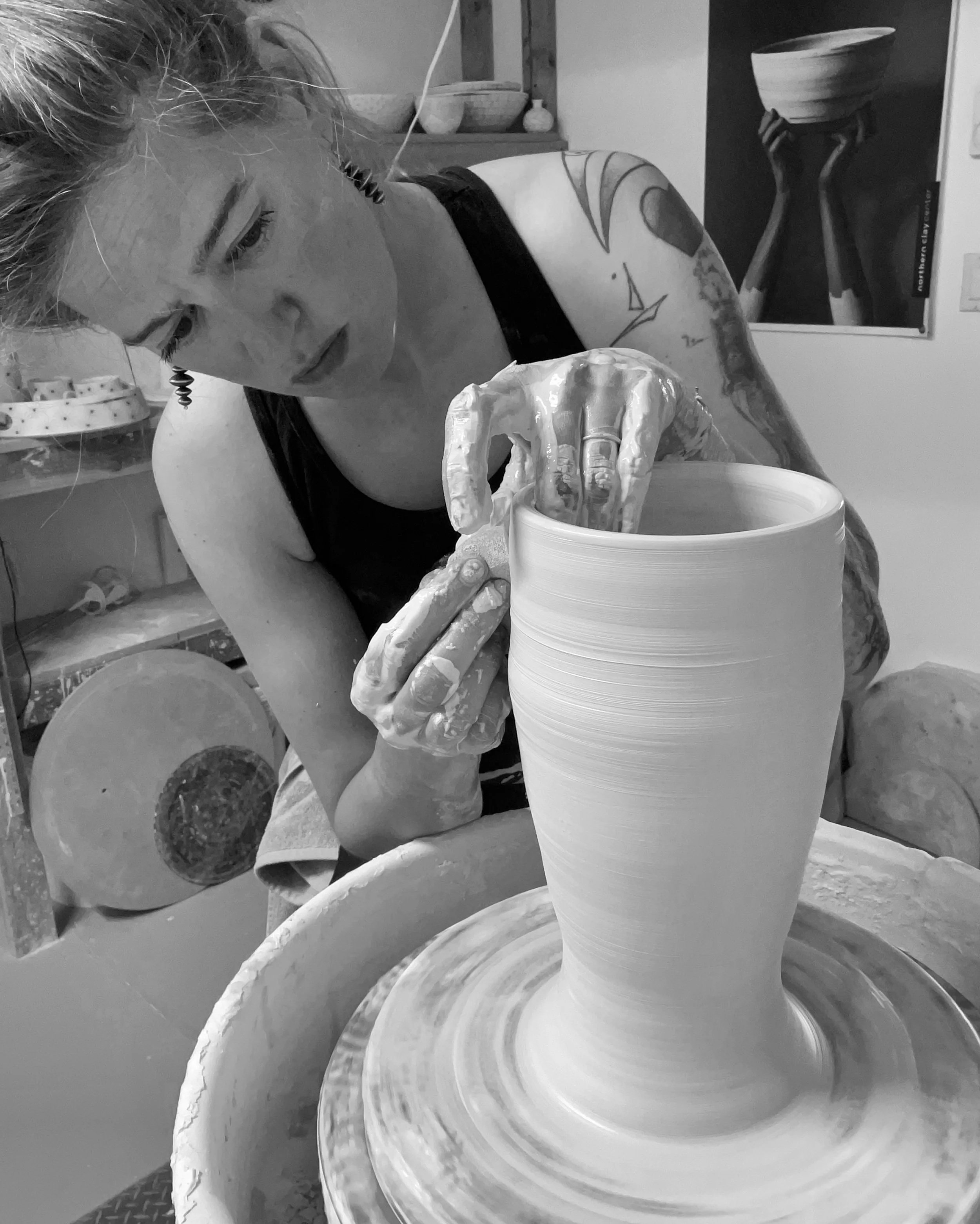 A woman with tattoos on her arm, focused on shaping a tall ceramic vase on a pottery wheel in a studio.