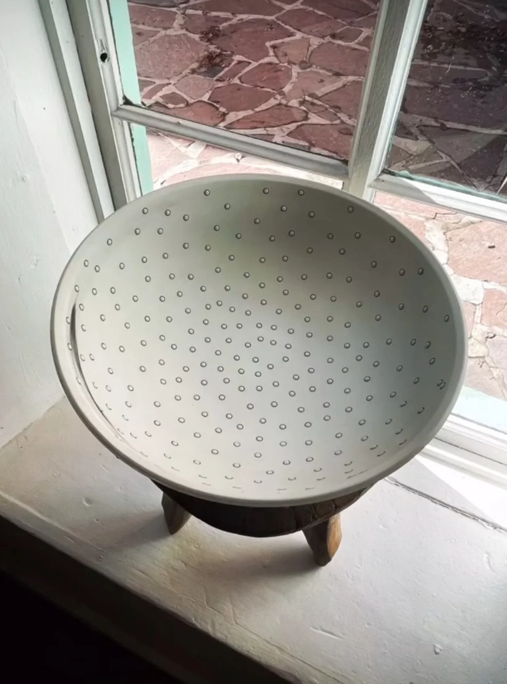 A white metal bowl with small holes, placed on a wooden stand, next to a window with a stone patio outside.
