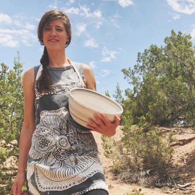 Young woman standing outdoors on a sunny day, holding a ceramic bowl, wearing a black and white patterned dress, with trees and blue sky in the background.