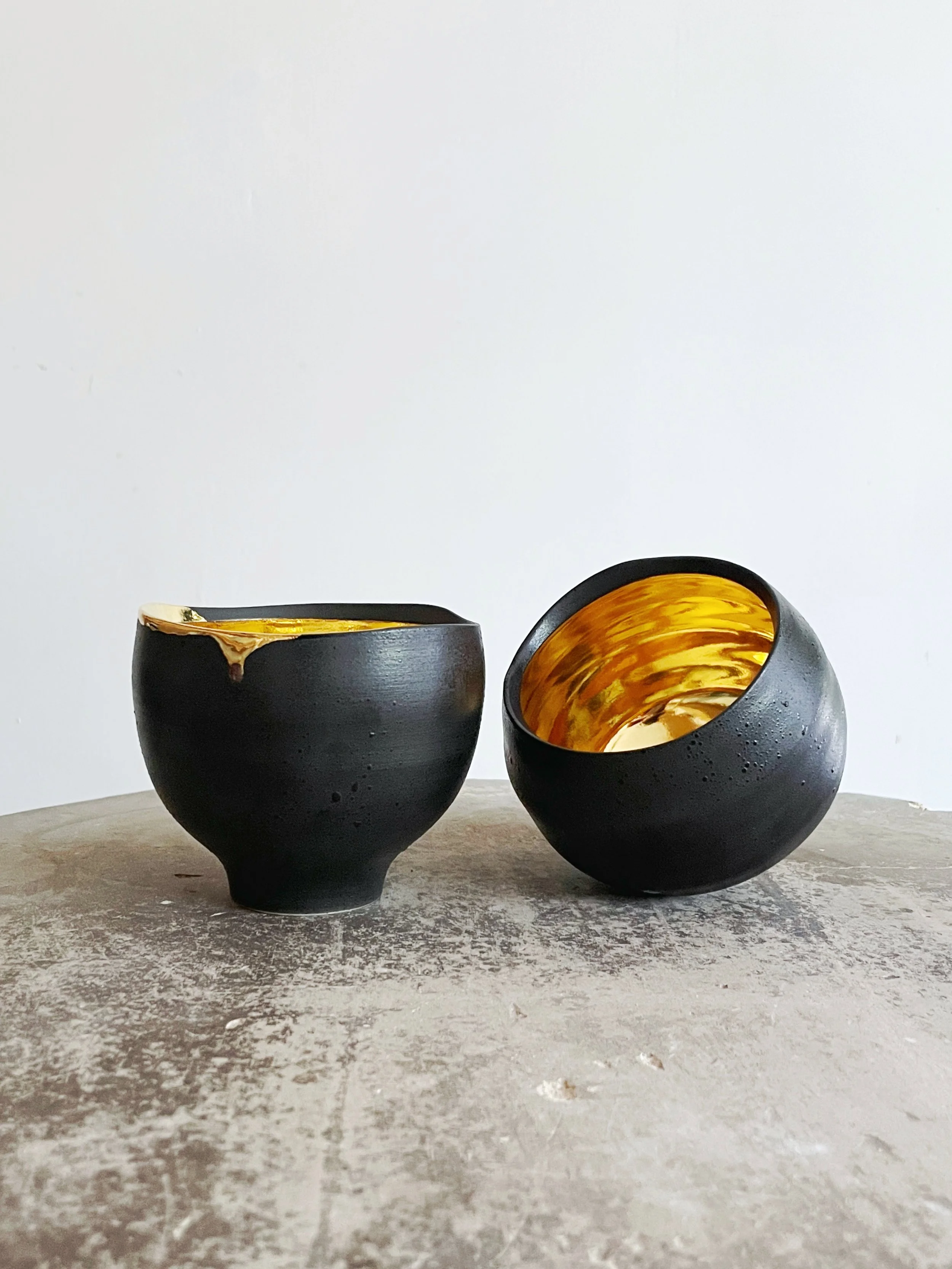 Two black and gold ceramic bowls placed on a rough concrete surface, with a plain white wall in the background.