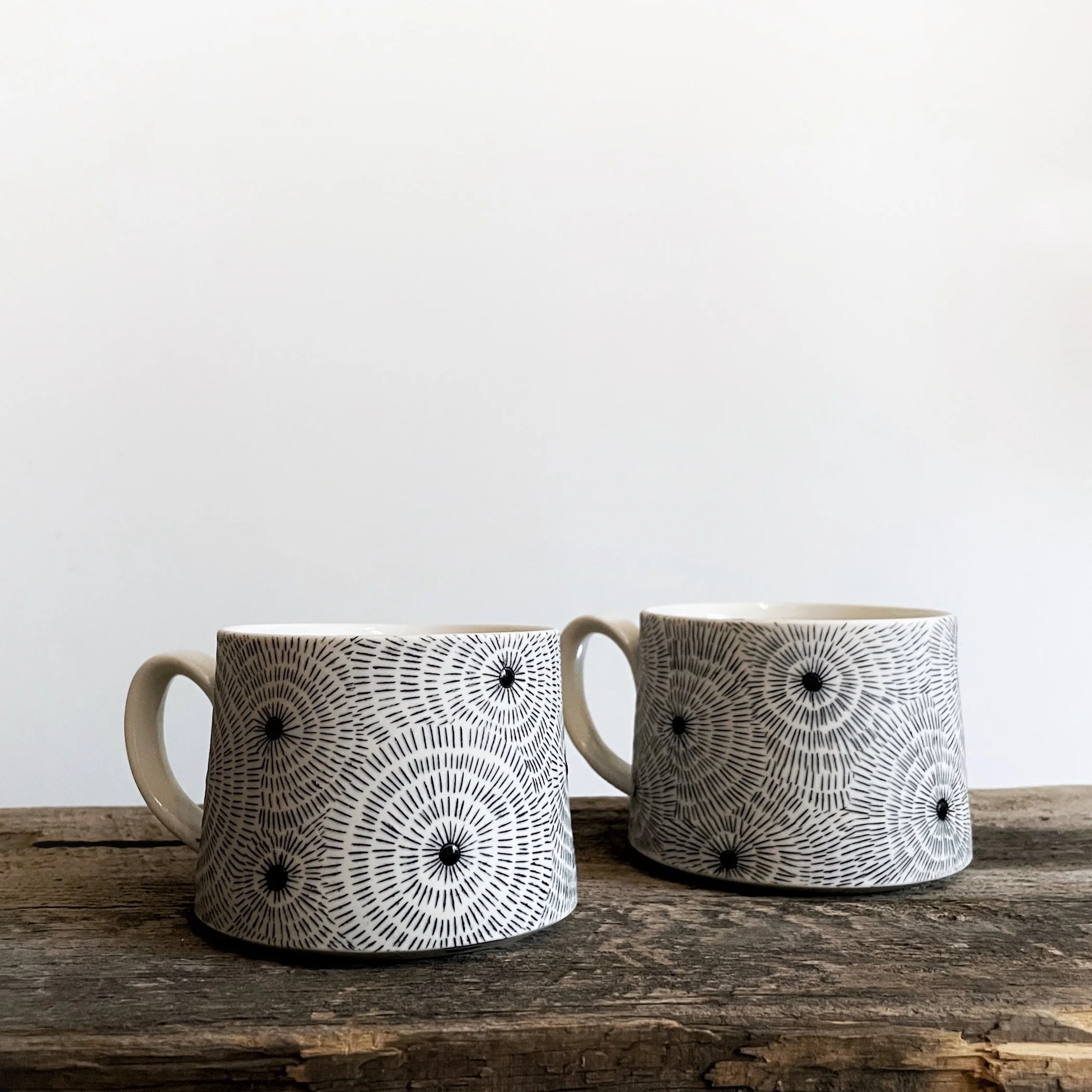 Two white ceramic mugs with black geometric patterns placed on a rustic wooden surface against a plain white background.