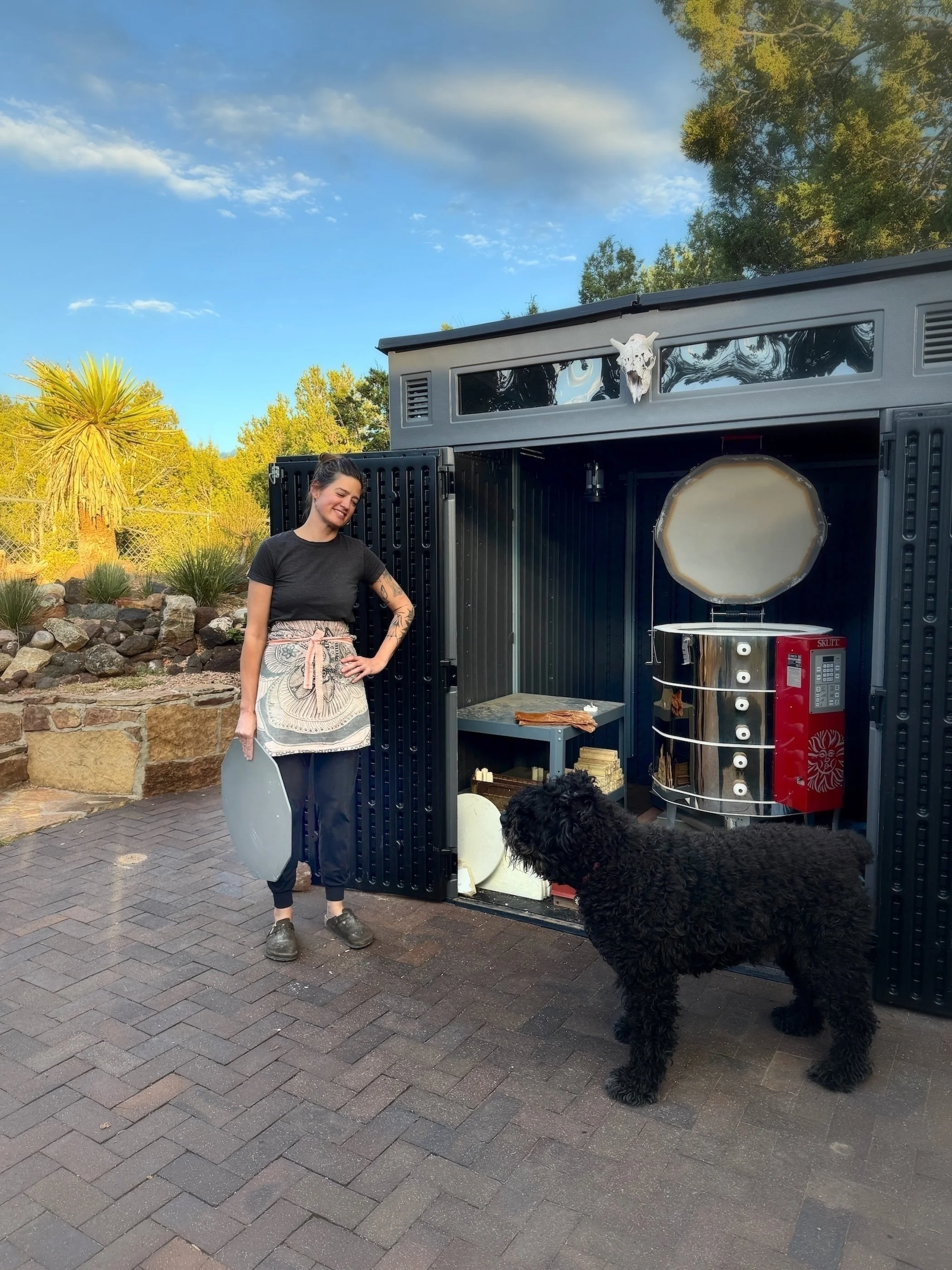 A woman smiling next to a large black outdoor oven with a dog standing nearby, surrounded by trees and a stone wall in the background.