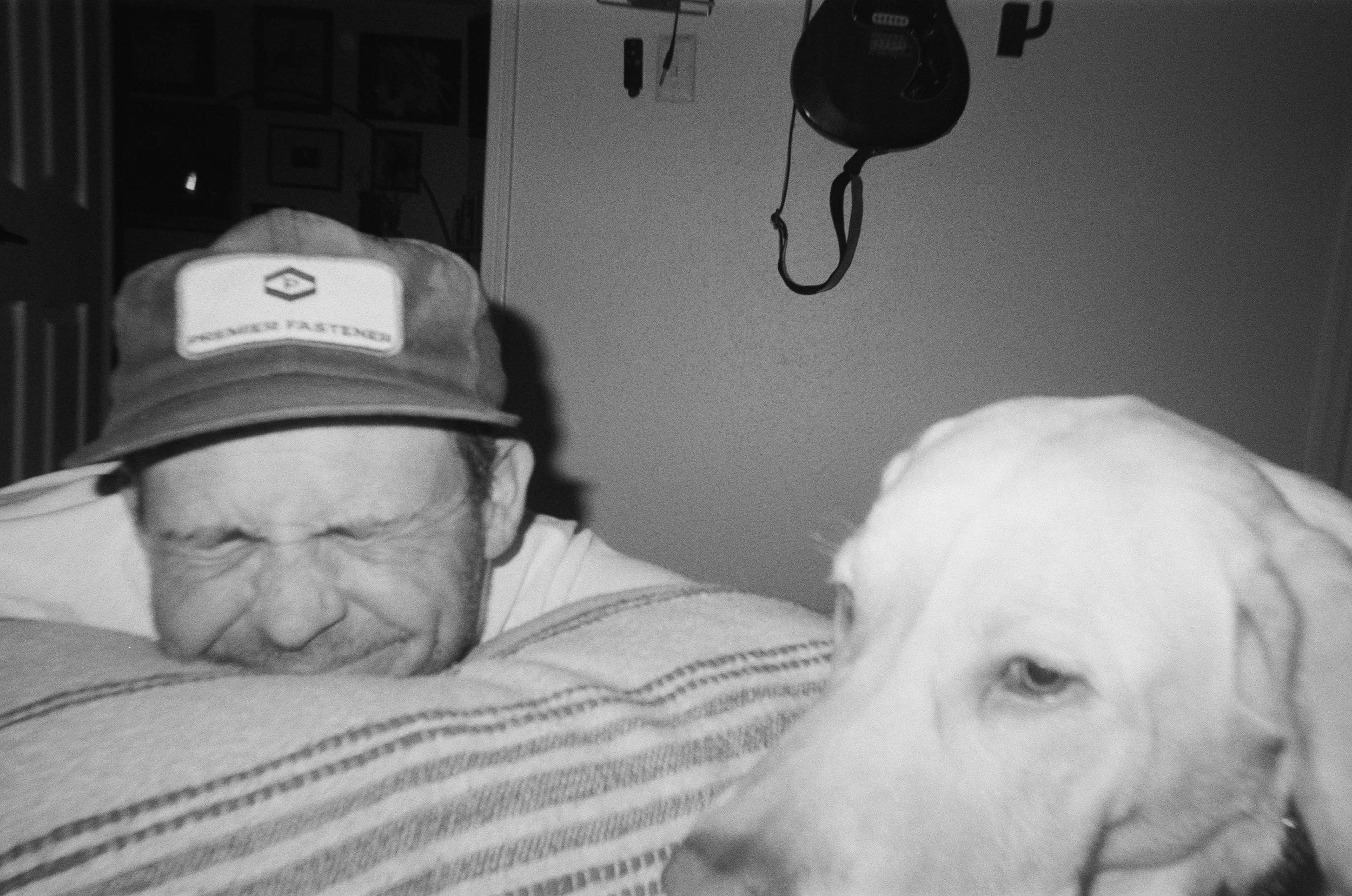 A person with a hat and glasses leaning on a striped pillow, next to a dog with light-colored fur, seen in a black-and-white photo.