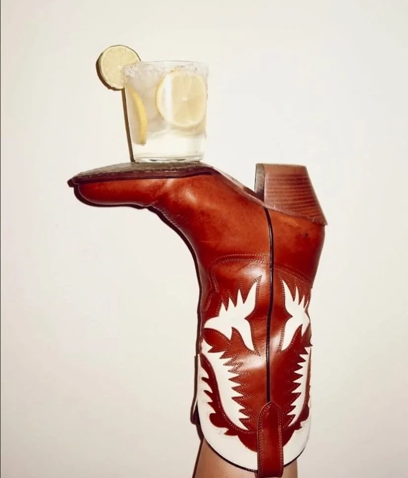 A brown leather cowboy boot with white decorative stitching, holding a glass of water with lemon slices on a plain background.