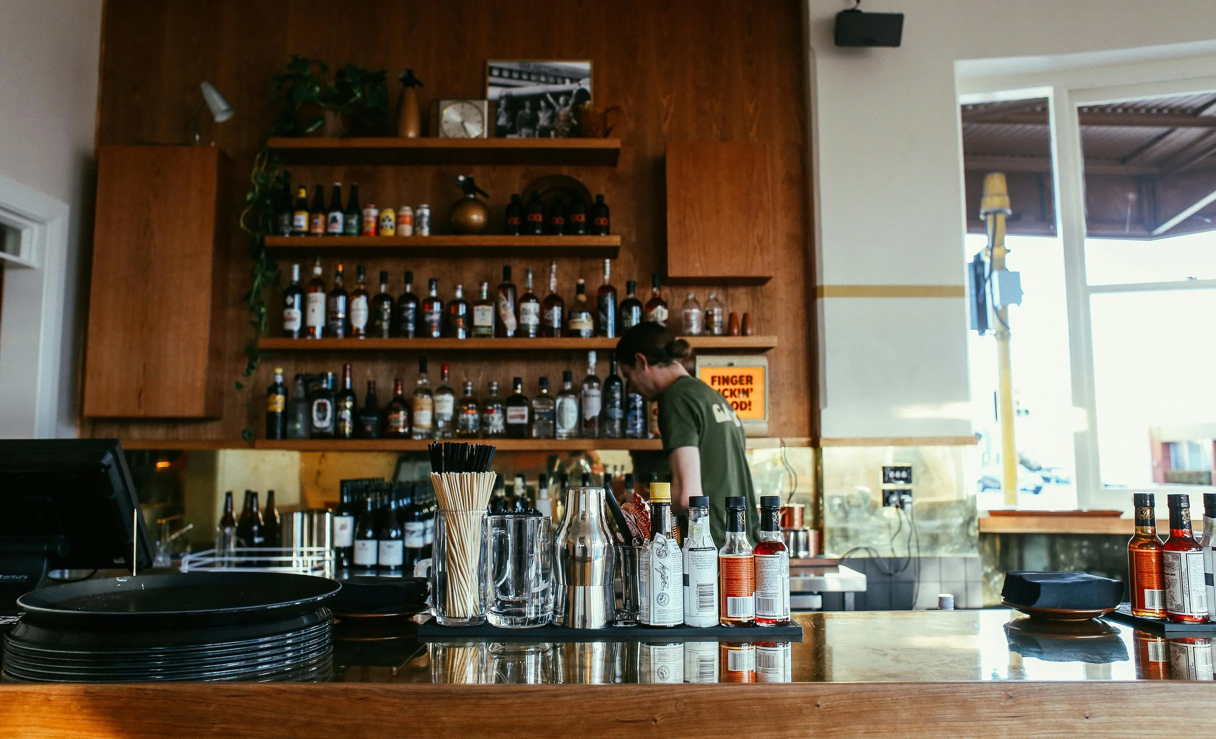 A bar or restaurant interior featuring a wooden wall with shelves holding bottles of alcohol and decorative items. There is a person working behind the bar, with various bottles, glasses, and utensils on the counter. A large window allows natural light to illuminate the space.