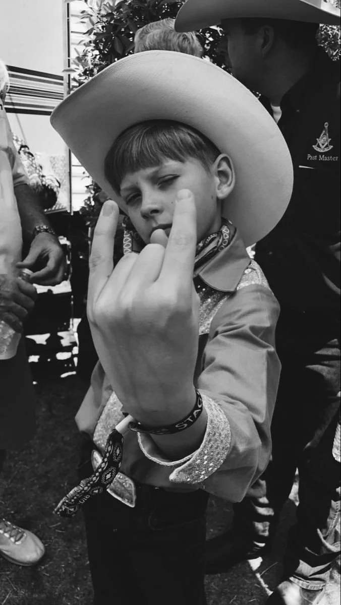 A young boy wearing a cowboy hat and a bandana around his neck making a rebellious gesture with his middle fingers, standing among others.