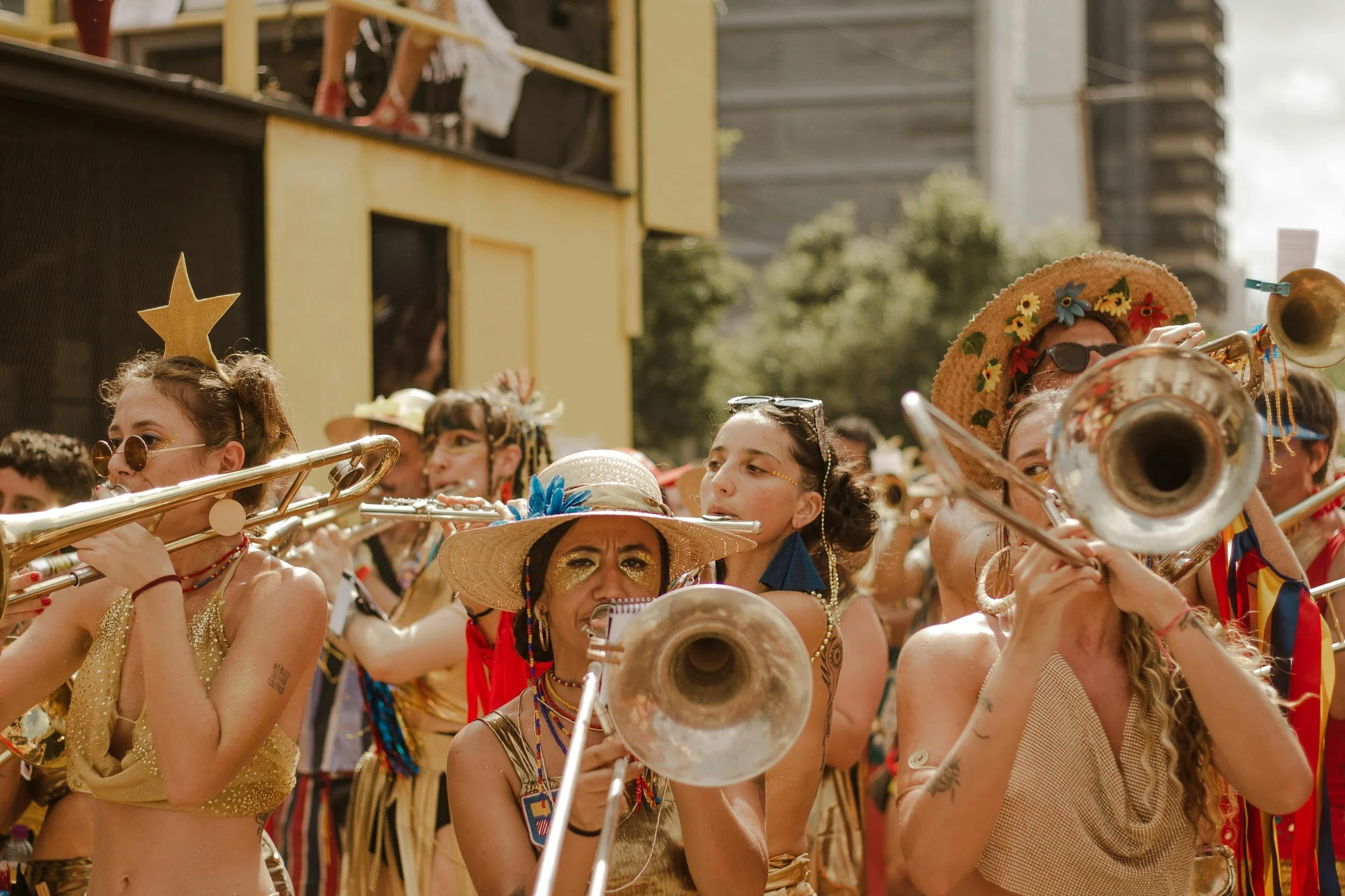 Group of women playing brass instruments during outdoor parade, dressed in colorful, festival attire with accessories like hats, sunglasses, and face paint.