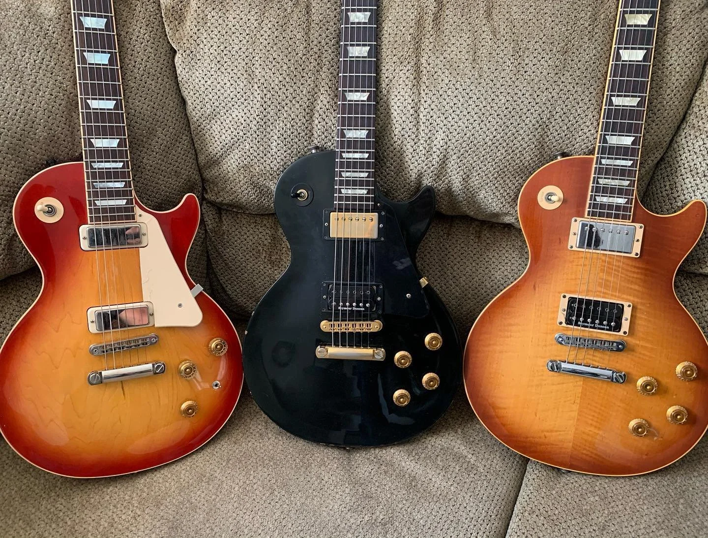Three electric guitars placed on a textured fabric sofa, from left to right: a sunburst Gibson Les Paul Double Cutaway, a black Gibson Les Paul Custom, and a natural finish Gibson Les Paul Standard.