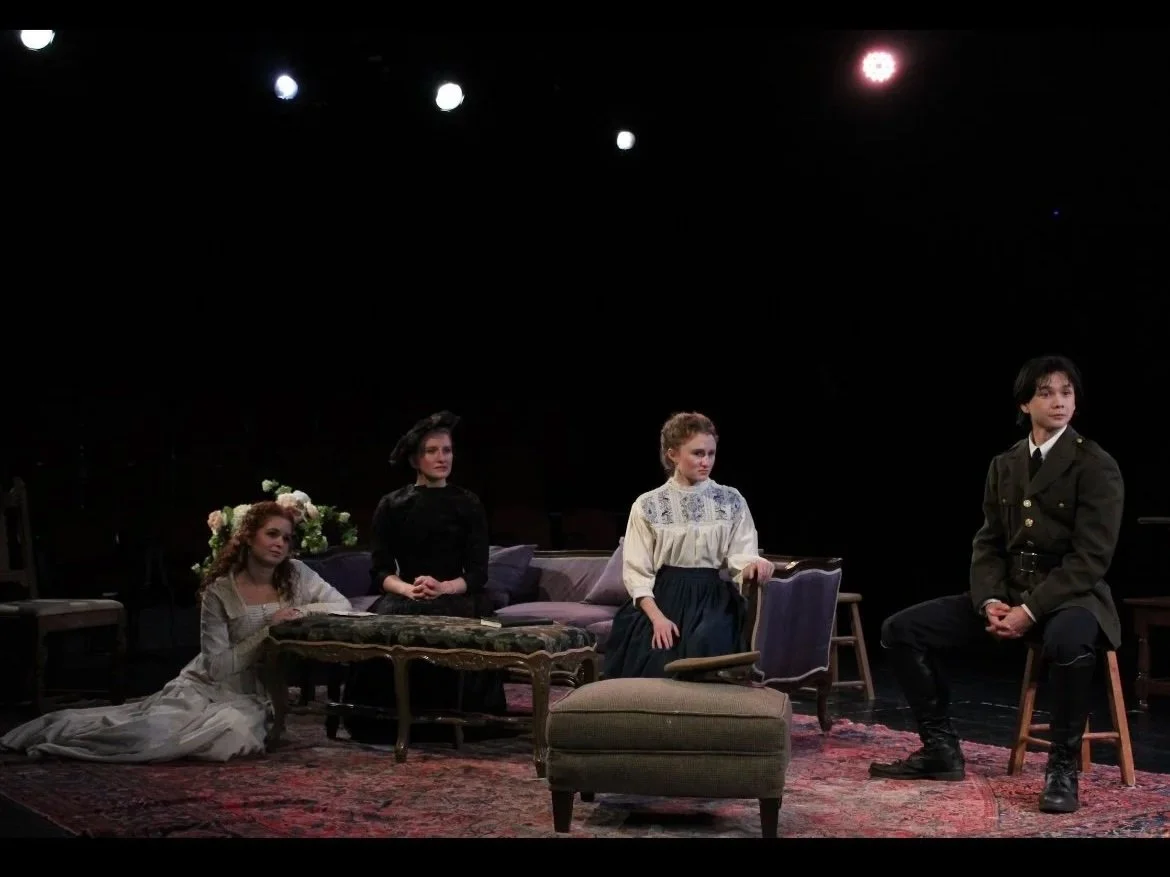 Four women and one man on a stage set for a play, with furniture and flowers, dressed in period costumes, under stage lighting.