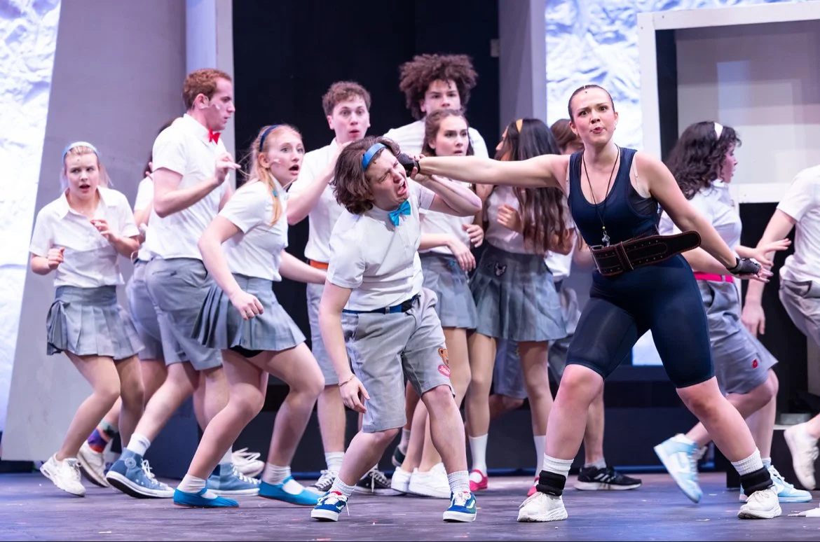 A stage scene with a woman yelling at a group of children dressed in school uniforms, some are surprised and scared while others look confused. The woman is pushing one child's head away.