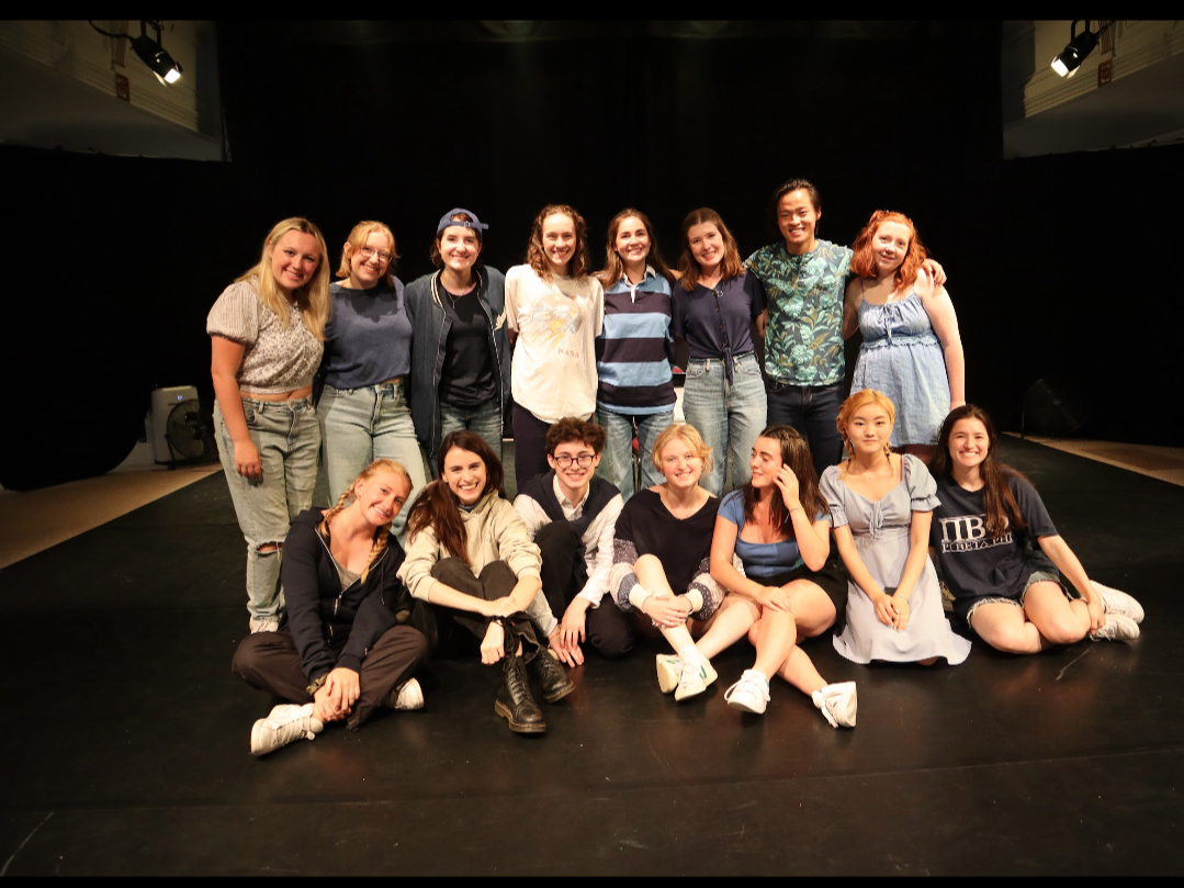 A group of fifteen young women and girls posing on a stage with a black backdrop in a theater or studio setting. They are smiling and standing or sitting in two rows, with some standing at the back and others sitting in front.