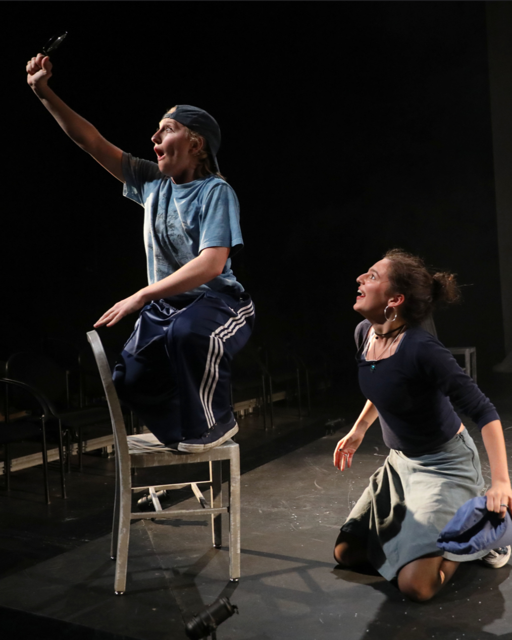 Two women on stage, one kneeling and the other sitting on a chair, with the woman on the chair holding a knife and looking up, suggesting a theatrical performance or play.