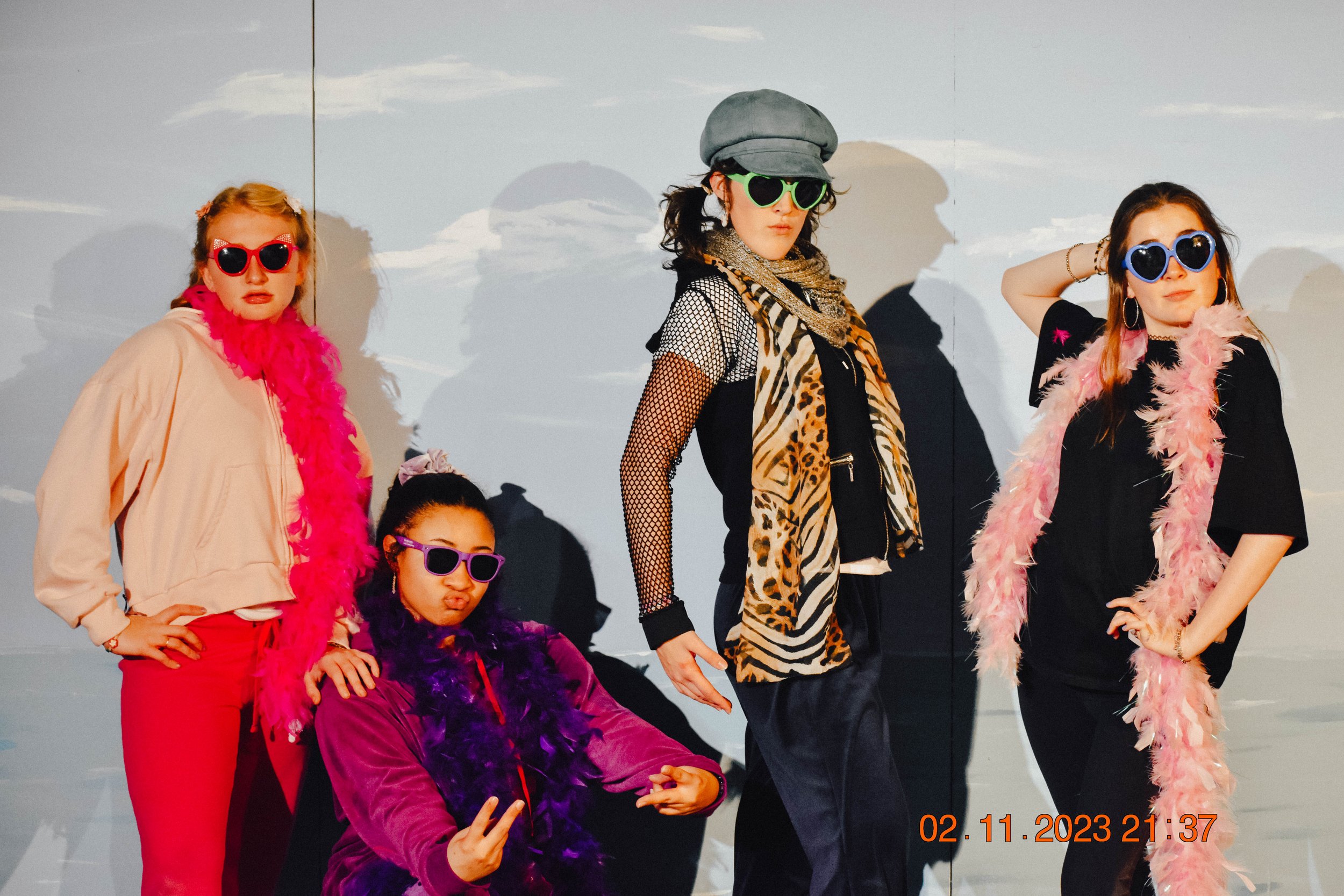 Four women dressed in colorful, playful outfits with feather boas and sunglasses, posing in front of a painted backdrop of a sky with clouds.