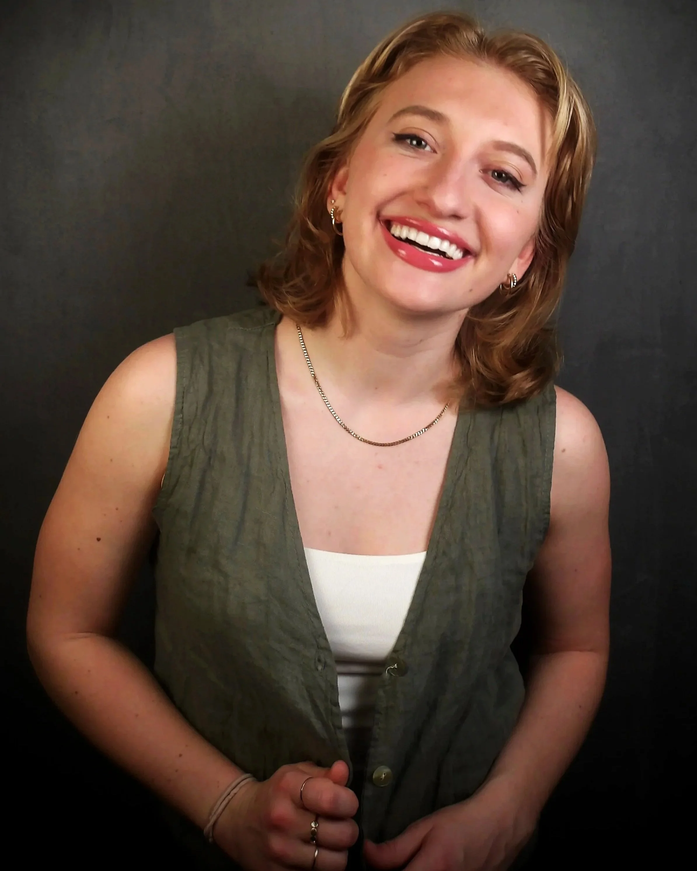 Portrait of a woman with shoulder-length reddish hair, smiling, wearing a sleeveless green vest over a white top, with jewelry including earrings, a necklace, and rings, standing against a dark background.