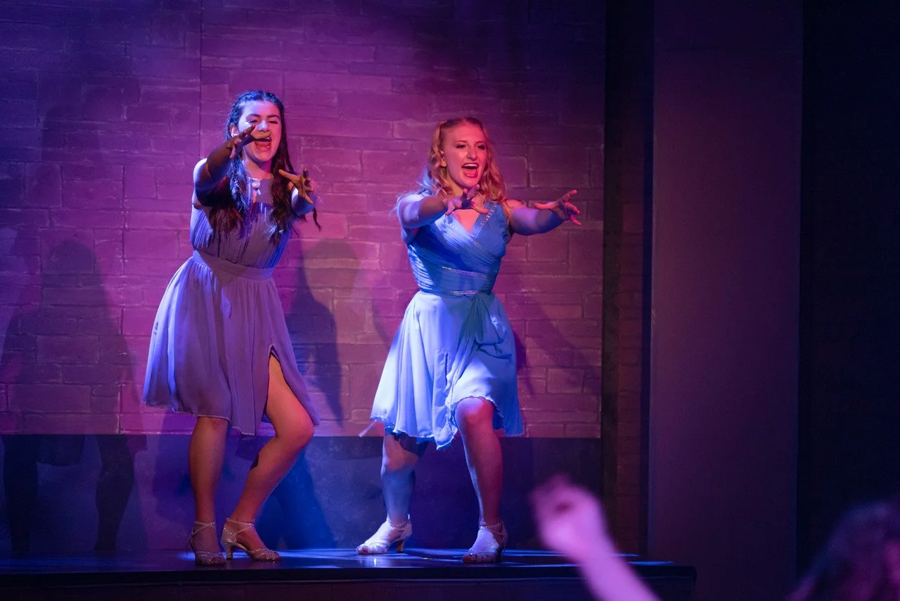 Two women in blue dresses performing on stage under purple lighting, with a brick wall background.