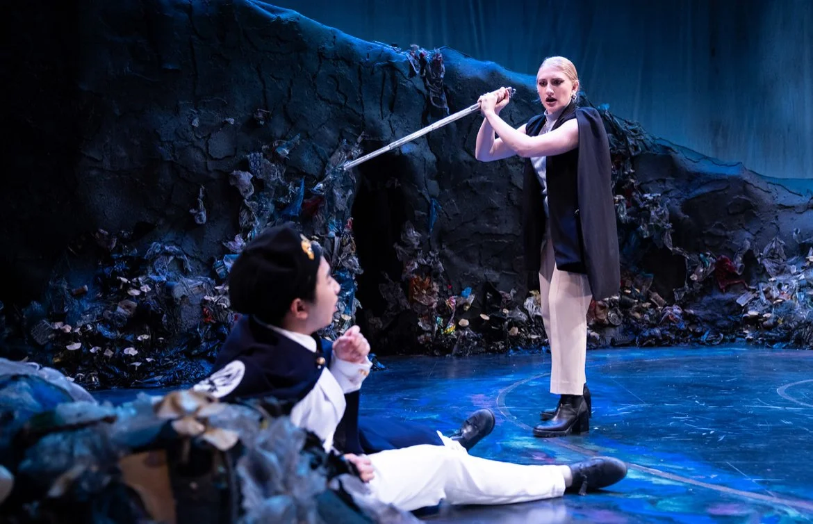 Actors on stage performing a scene with a dark, rocky backdrop and trash, with one actor standing holding a weapon and another actor sitting on the ground.