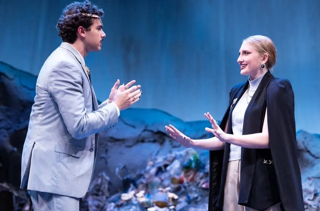 Two actors on stage, a man in a light-colored suit and a woman in a dark blazer and light blouse, engaged in conversation against a backdrop of a scene with garbage and trash.