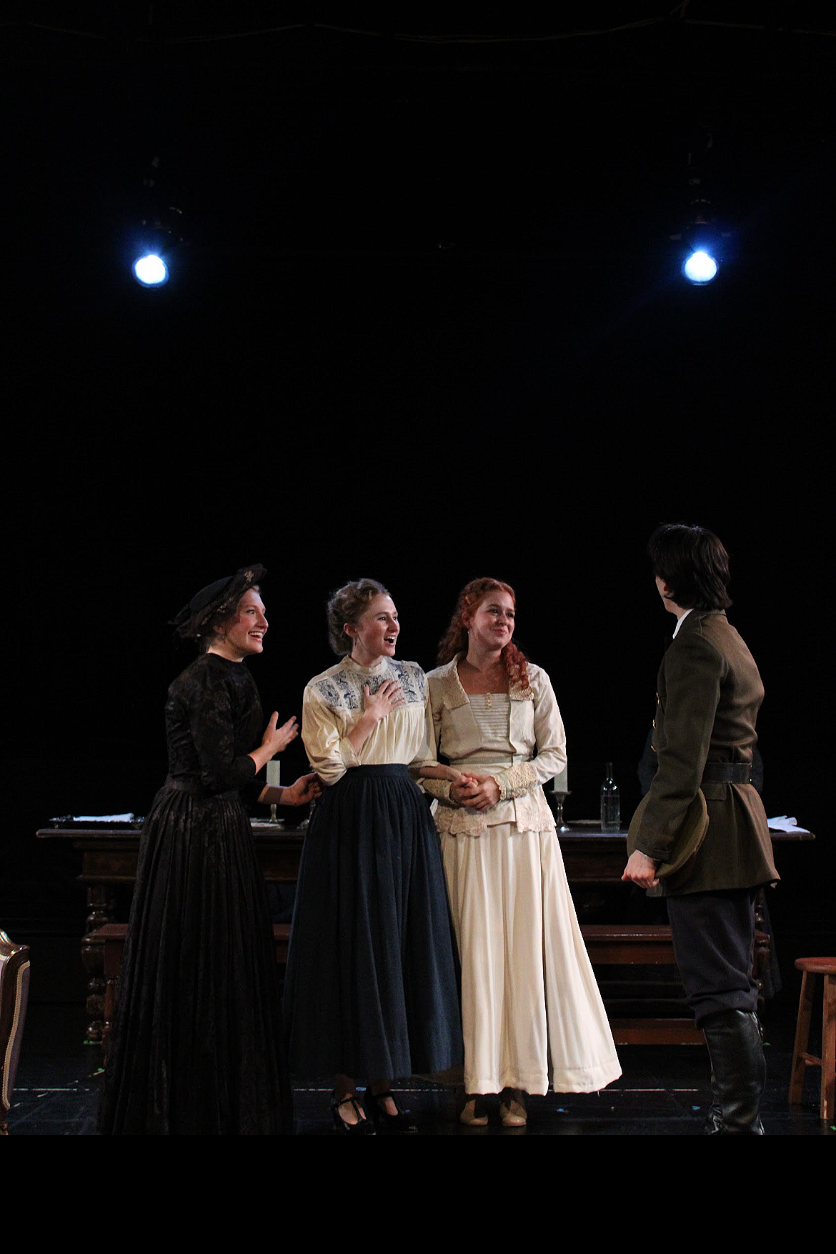 Four women and one man in period costumes on stage, engaging in a theatrical scene, with a dark background and stage lighting.
