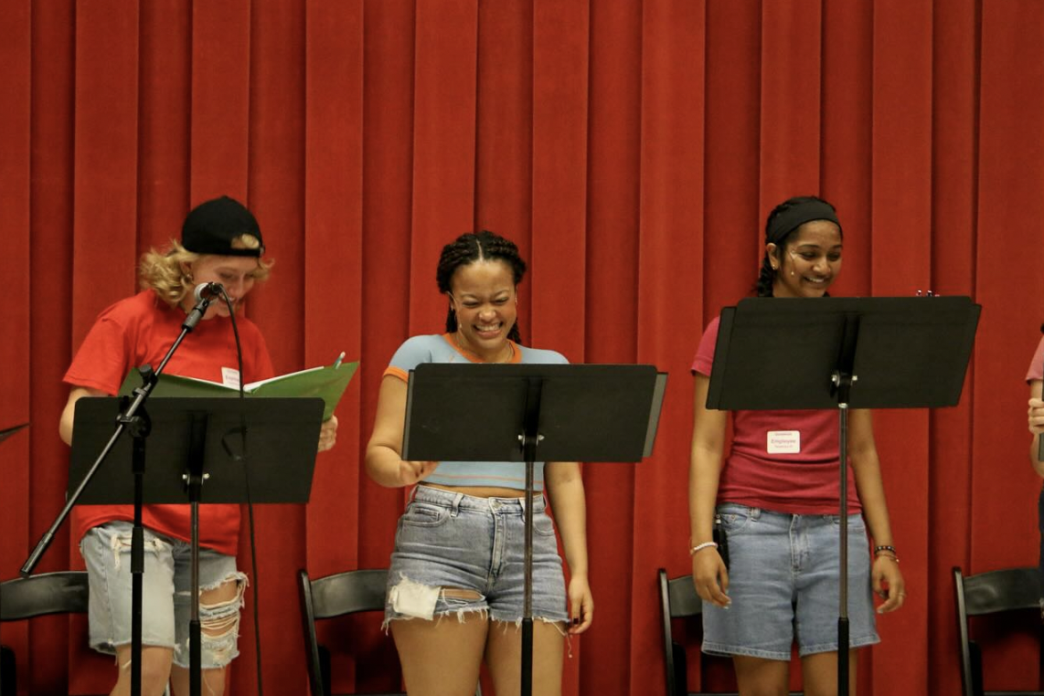 Three young women standing behind music stands, singing or performing on stage with a red curtain backdrop.
