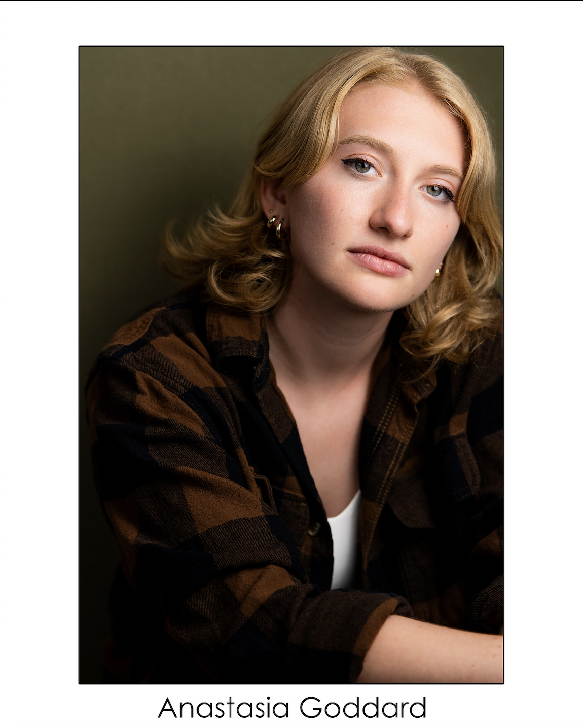 Portrait of a young woman with blonde hair, wearing a brown and black plaid shirt and earrings, against a dark green background.