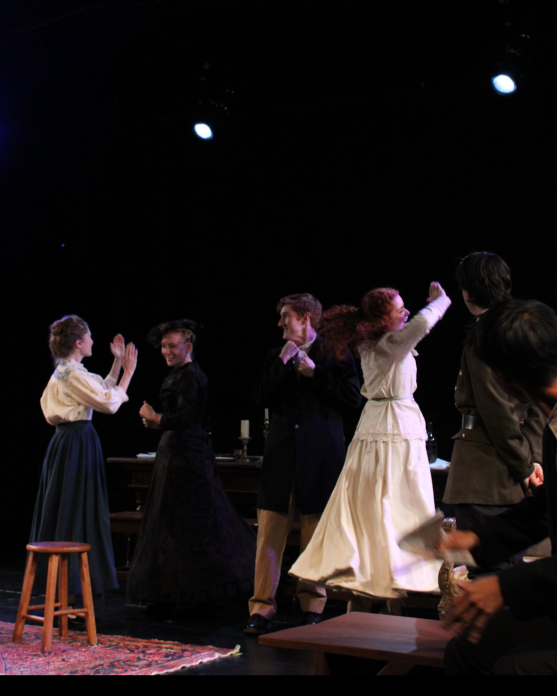 A group of actors on stage in period costumes, smiling and greeting each other during a theatrical performance.