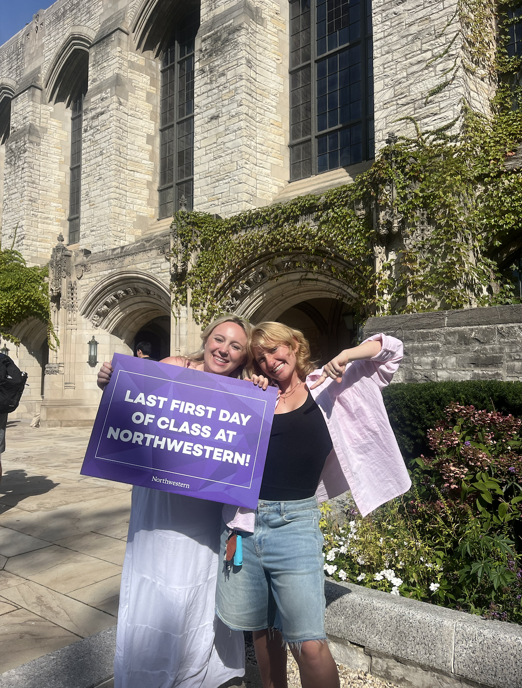 Two smiling young women standing outside a stone university building, holding a purple sign that reads 'Last First Day of Class at Northwestern!'