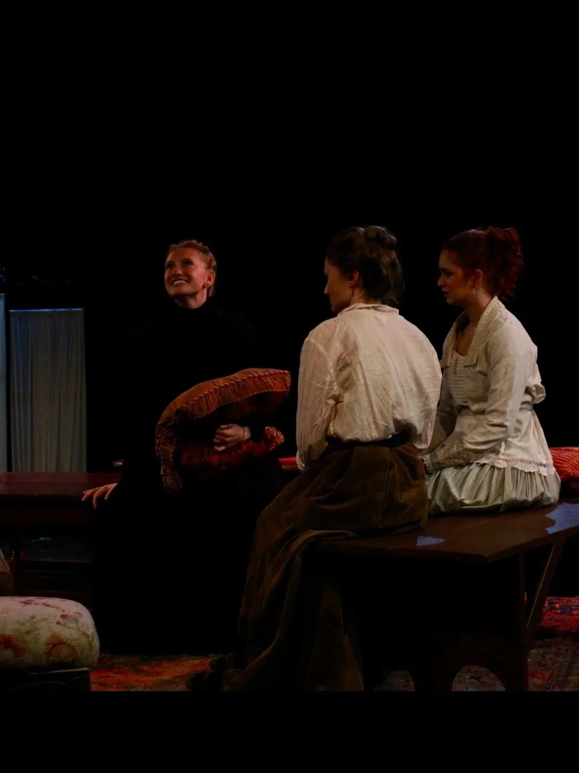 Three women in period costumes sitting on a low platform, with one woman standing nearby, in a theatrical performance.