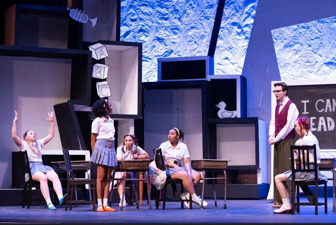Children in a school classroom play, with some sitting and others standing, and a man in a vest watching. The background has shelves with a duck illustration, a chalkboard with part of the phrase 'I CAN READ,' and decorative alphabet blocks.