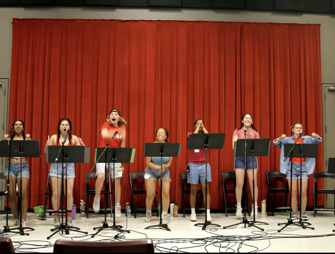 A group of seven young women standing on a stage with red curtains in the background, some singing into microphones, others clapping or covering their faces, and water bottles on the floor in front of them.