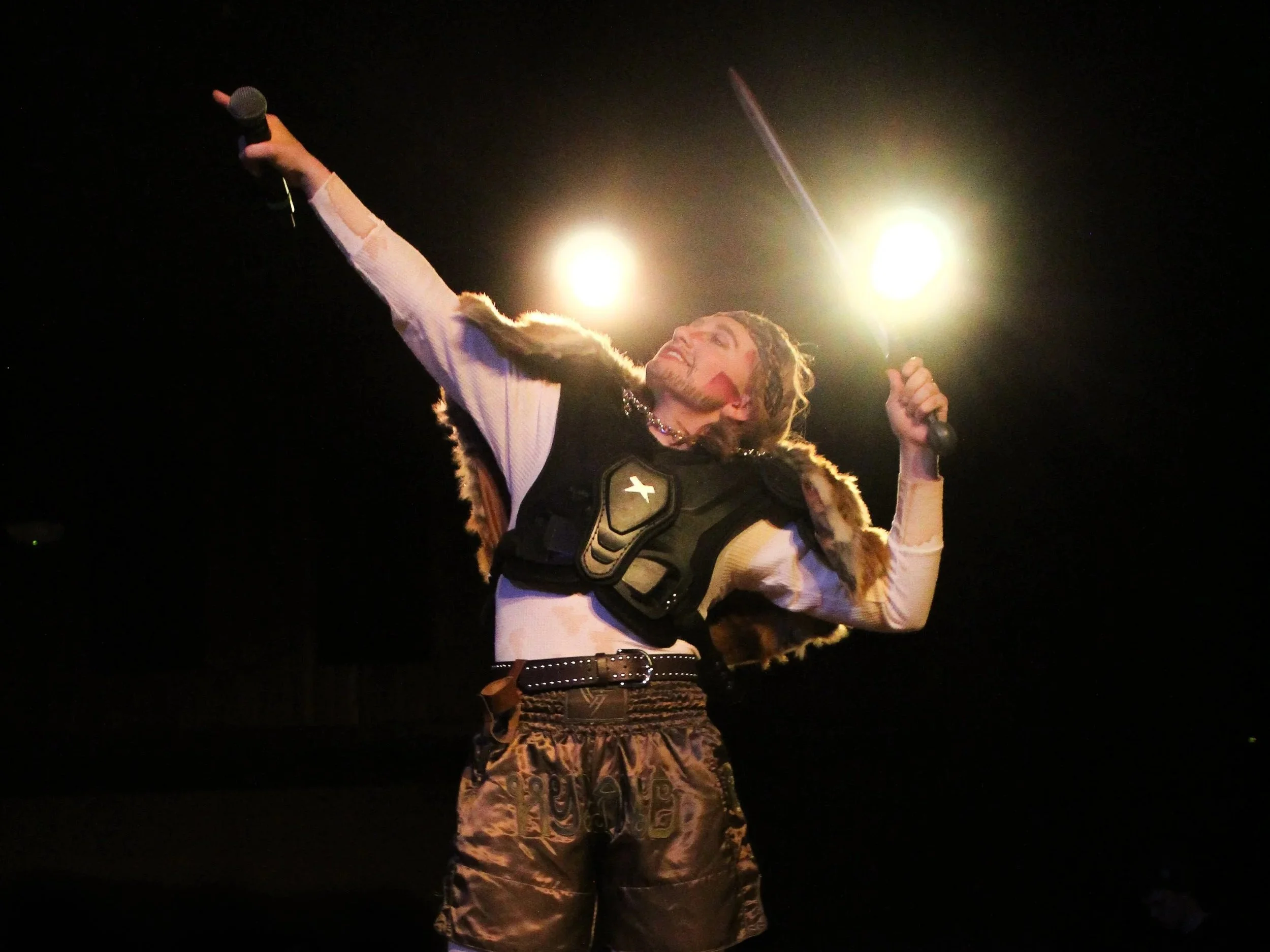 Performer on stage dressed in Viking costume, holding a microphone in one hand and a sword in the other, with bright stage lights behind.